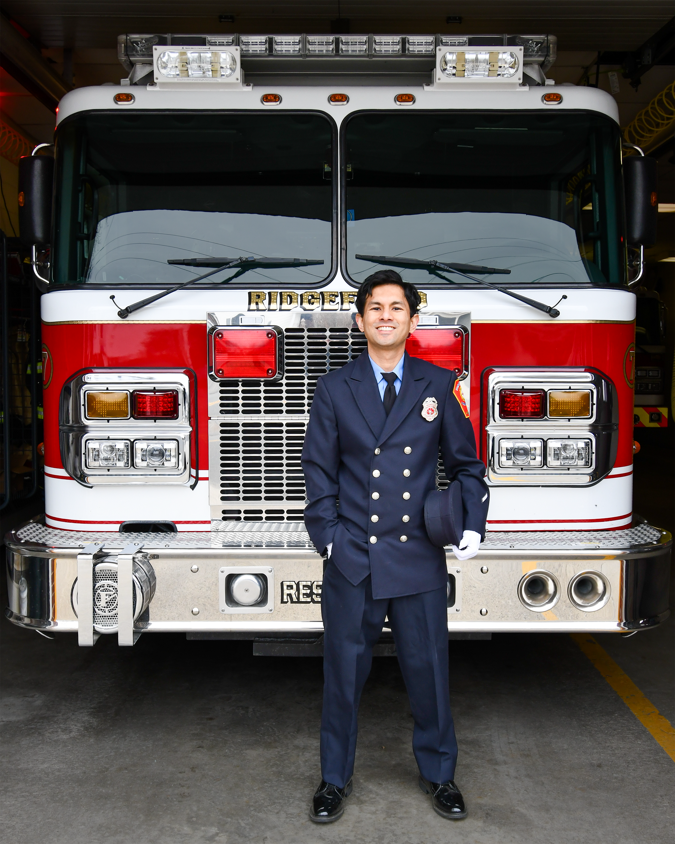 A firefighter standing in front of a fire truck inside a fire station, smiling, wearing a navy blue uniform and holding a peaked cap.