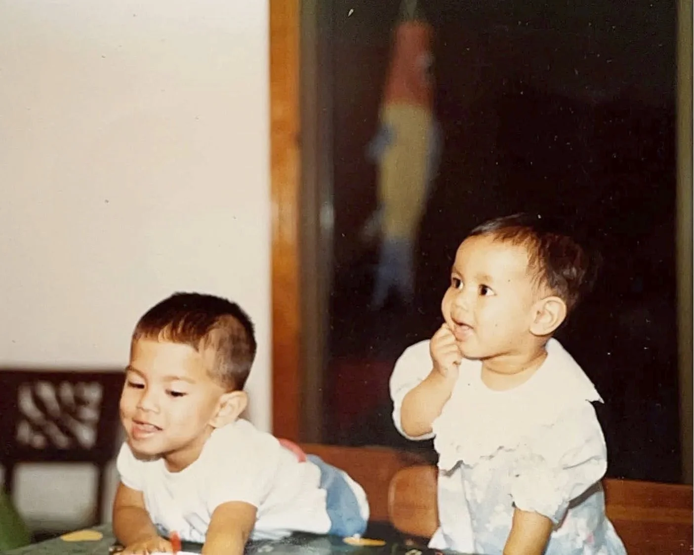 Two young children, a boy and a girl, are standing next to each other indoors. The boy is leaning forward on a table with his eyes closed and tongue slightly out, while the girl is looking to the right with a finger on her cheek, appearing curious or thoughtful.