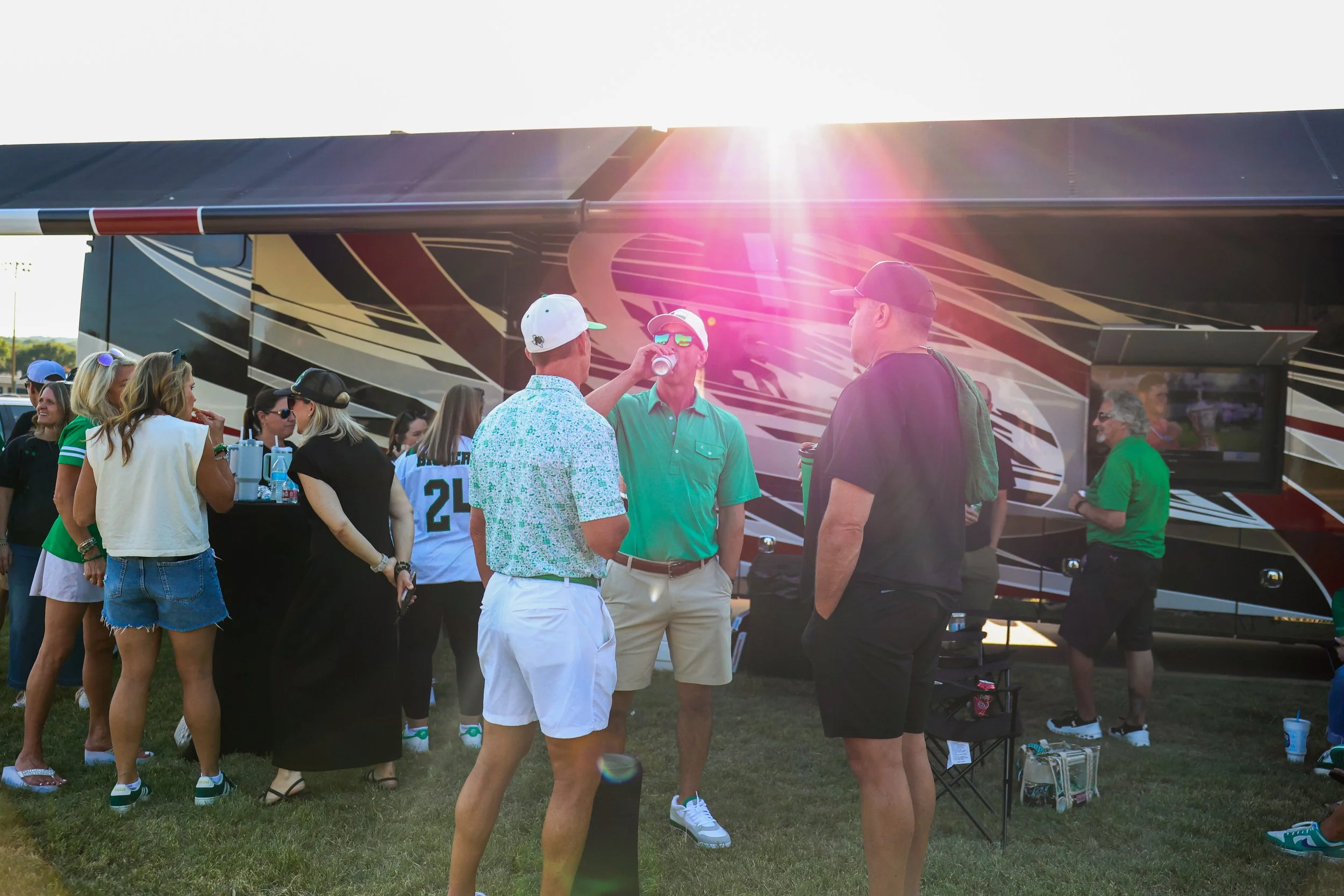Group of people at a social gathering outside with a large bus in the background, some talking and drinking, in casual summer attire.