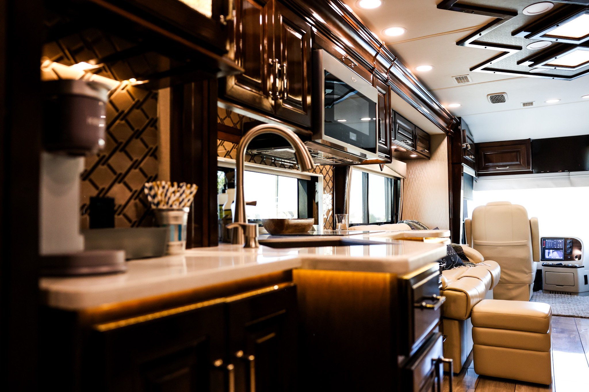 Interior of an RV with a kitchen area featuring dark wood cabinets, a microwave, a sink, and a seating area with a cream-colored chair and ottoman, and large windows in the background.