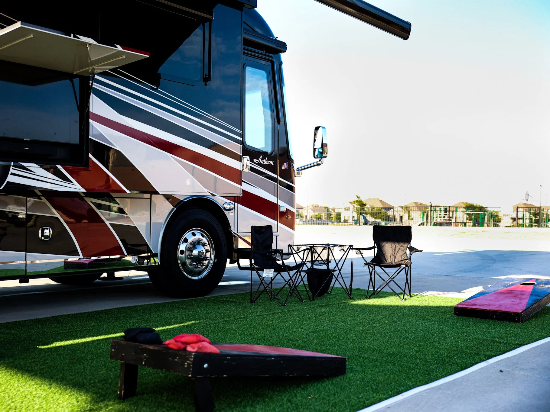 Outdoor scene with a black, white, and red recreational vehicle (RV) parked on a paved lot, next to artificial grass. There are four black camping chairs, a small foldable table with items, two cornhole boards, and a bright red skate board nearby. In the background, a fence and some houses are visible.