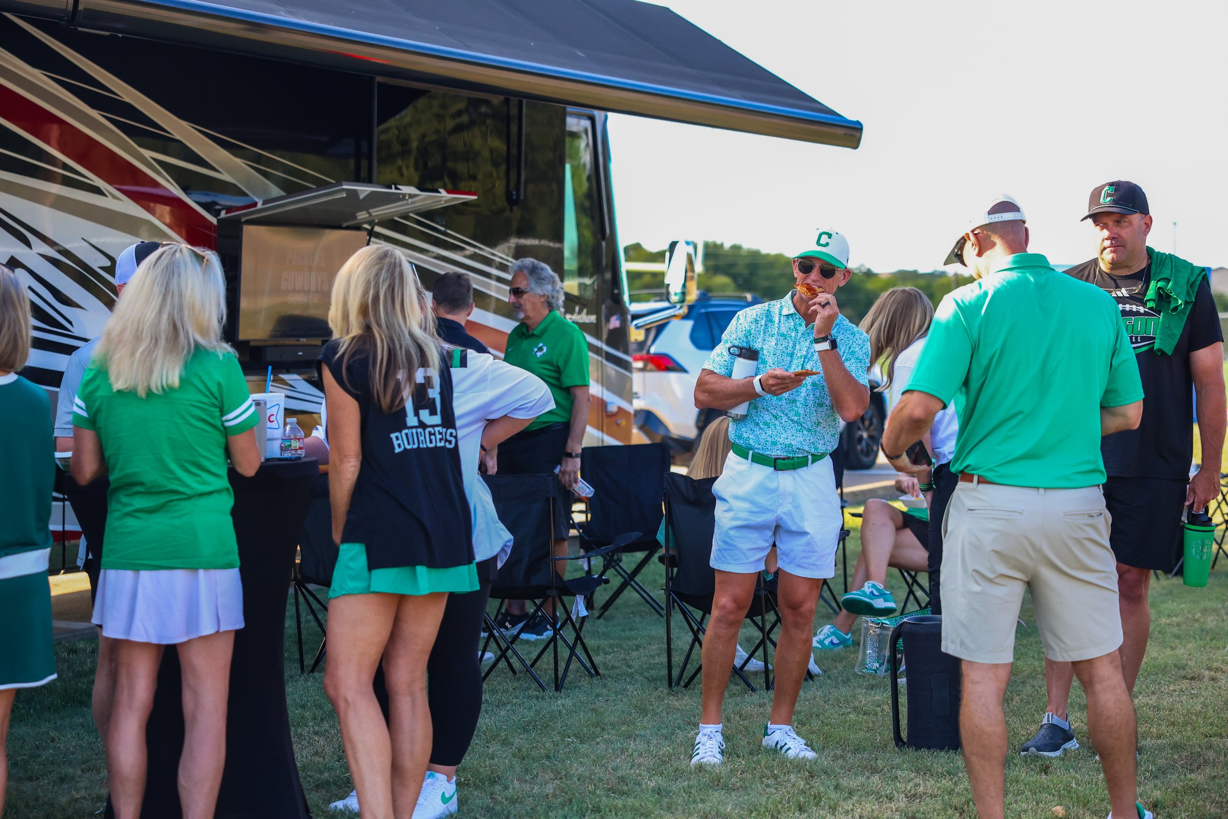 Group of people in sports attire gathered outdoors near a large bus, some eating and talking, with a woman in the foreground wearing a green shirt and white skirt, and a man in the center wearing a floral shirt, white shorts, green belt, and sunglass