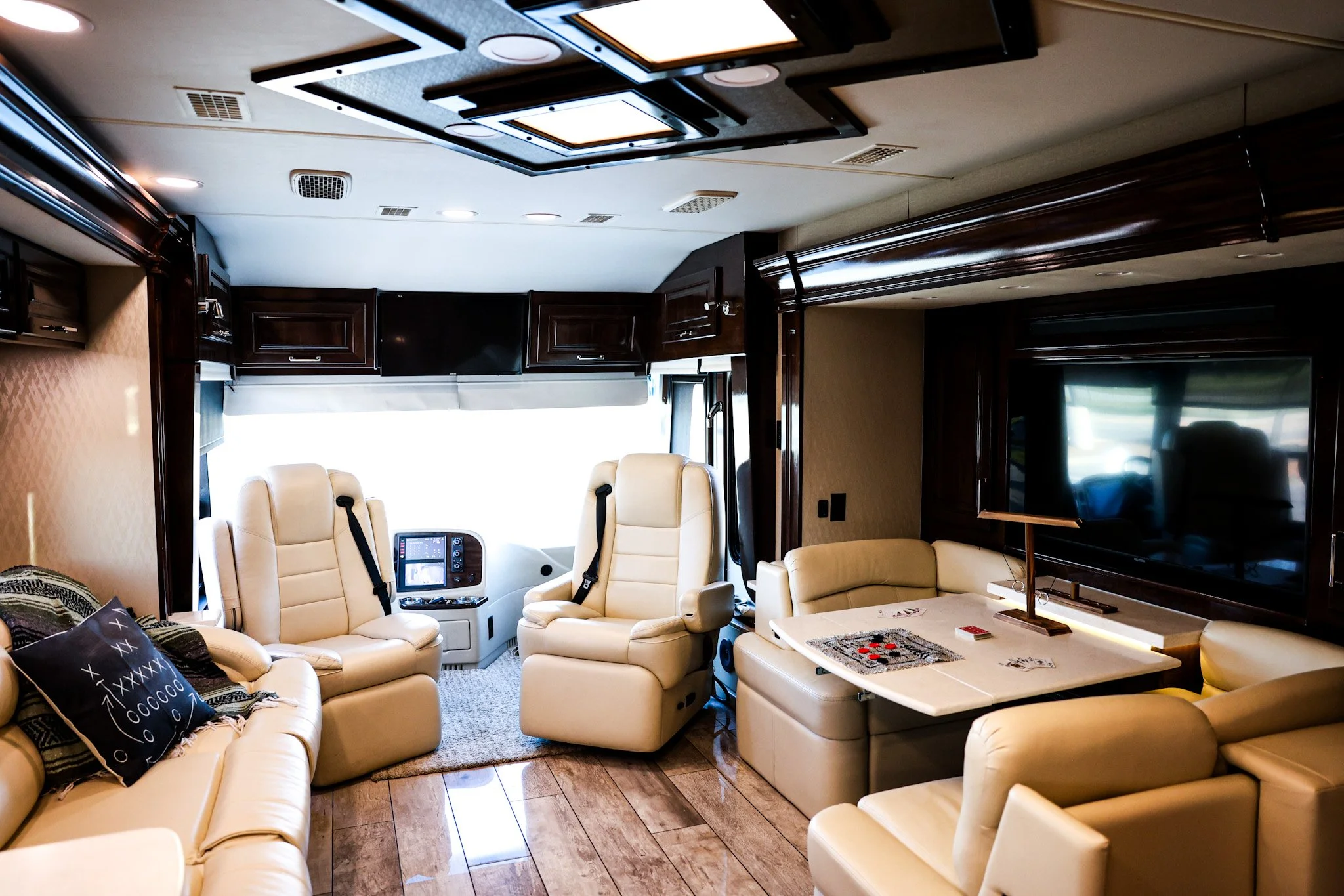 Interior of an RV with beige leather seating, a large flat-screen TV, a white table with games, and two captain's chairs at the front facing a large window.