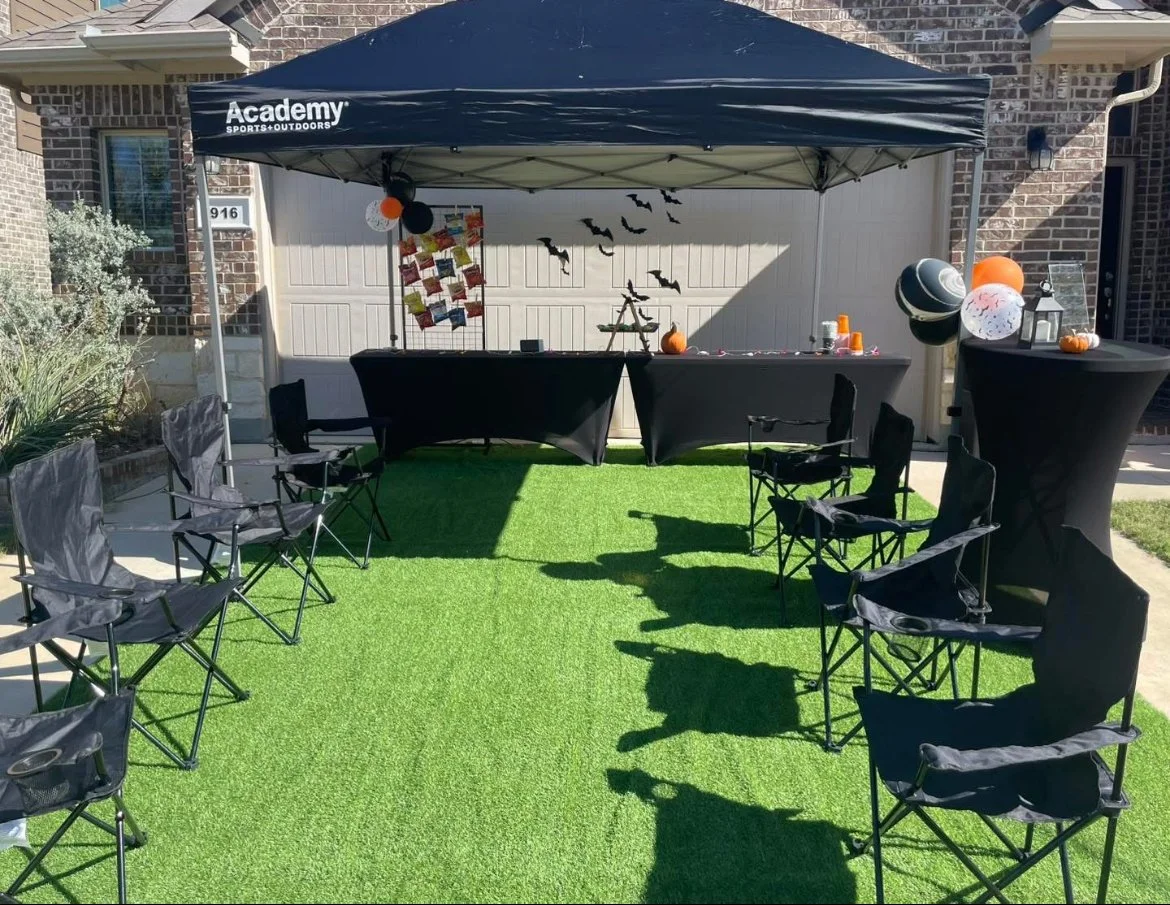 Halloween-themed outdoor setup with black tables, chairs, balloons, and decorations on artificial grass in front of a house garage, with bat and pumpkin decorations.