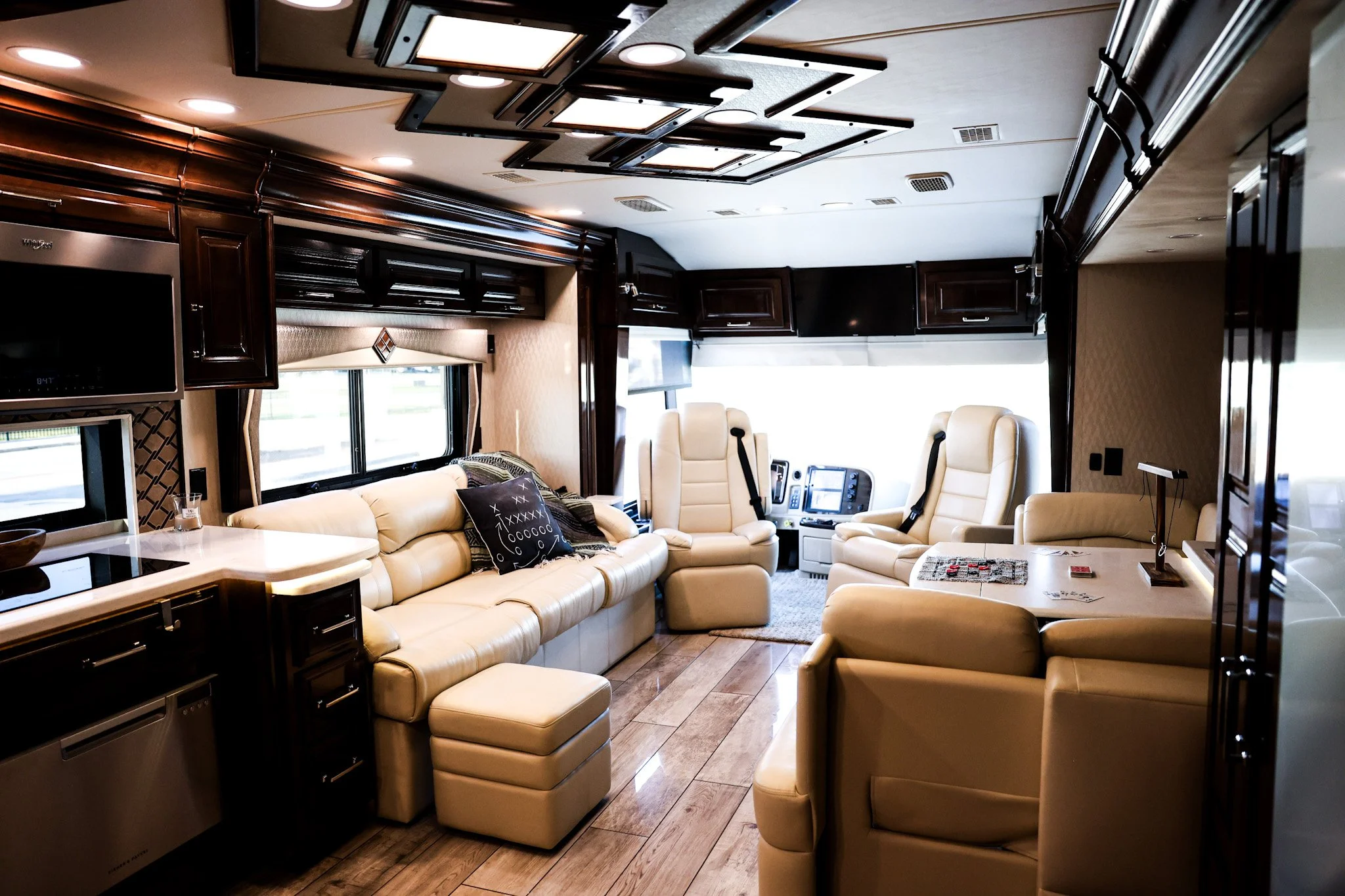 Interior of a luxurious RV with cream-colored leather seating, dark wooden cabinets, large windows, and a wood floor. The views include a kitchen area and a driving area at the front.