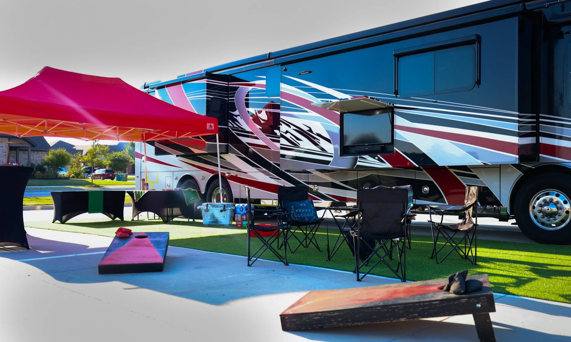 A large motocross racing truck parked on a driveway with outdoor chairs and tables under a red canopy tent, with a grassy area, houses, and trees in the background.