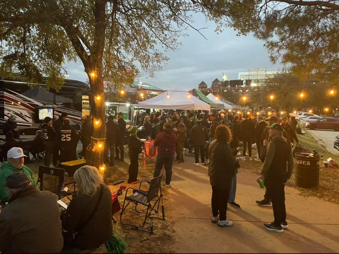 People gathering outdoors at a nighttime tailgate party with string lights, tents, and RVs in a parking lot.