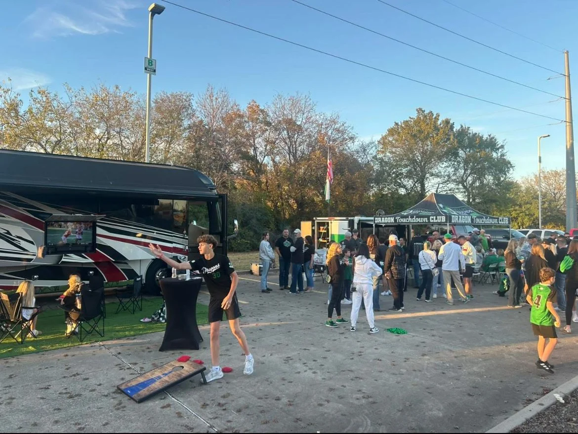 People gather at an outdoor event with a large bus and food trucks, some children playing bean bag toss, in a parking lot surrounded by trees and streetlights.