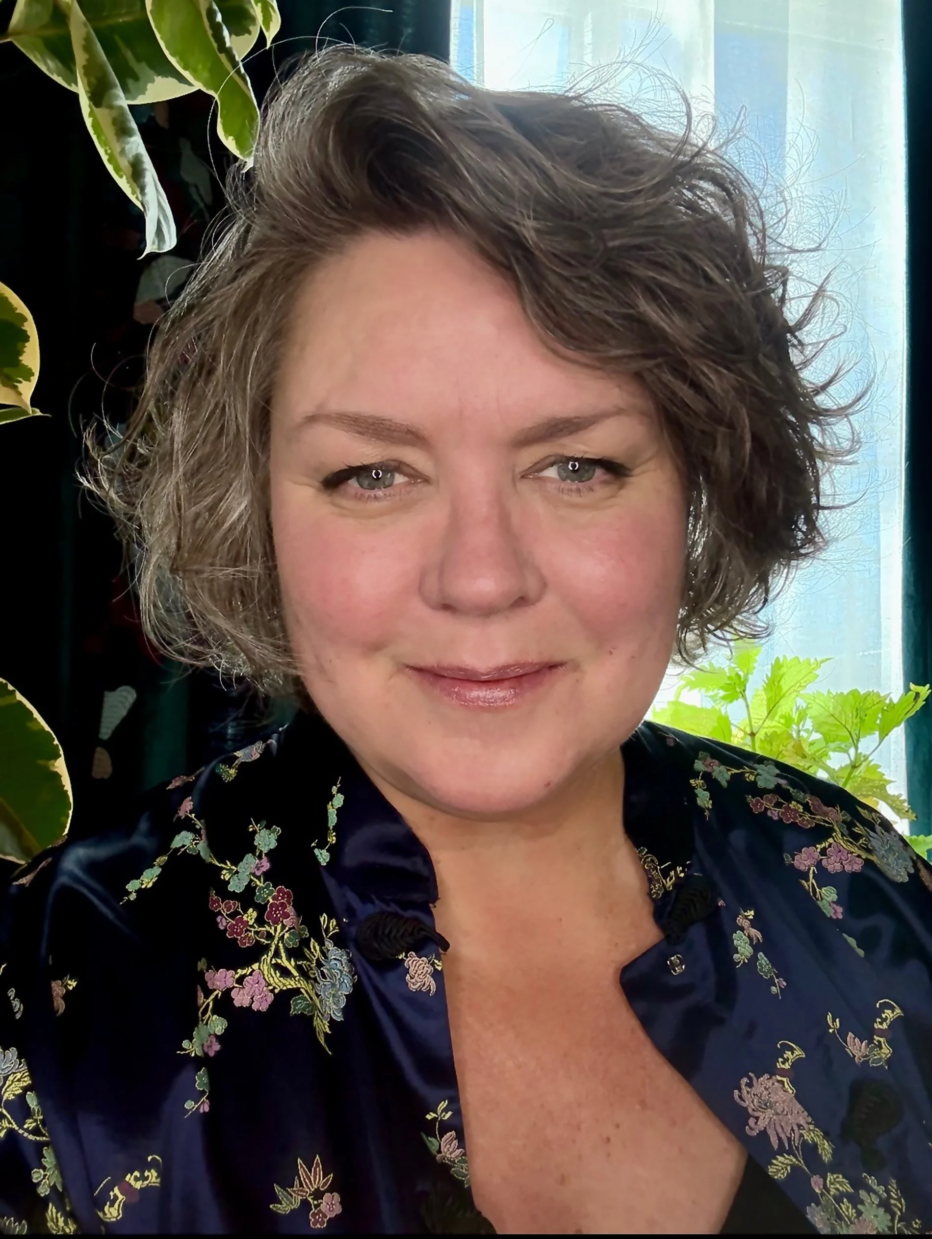 A close-up portrait of a woman with short, curly brown hair, blue eyes, and fair skin, smiling gently. She is wearing a dark satin jacket with floral embroidery, standing in front of green plants near a window.