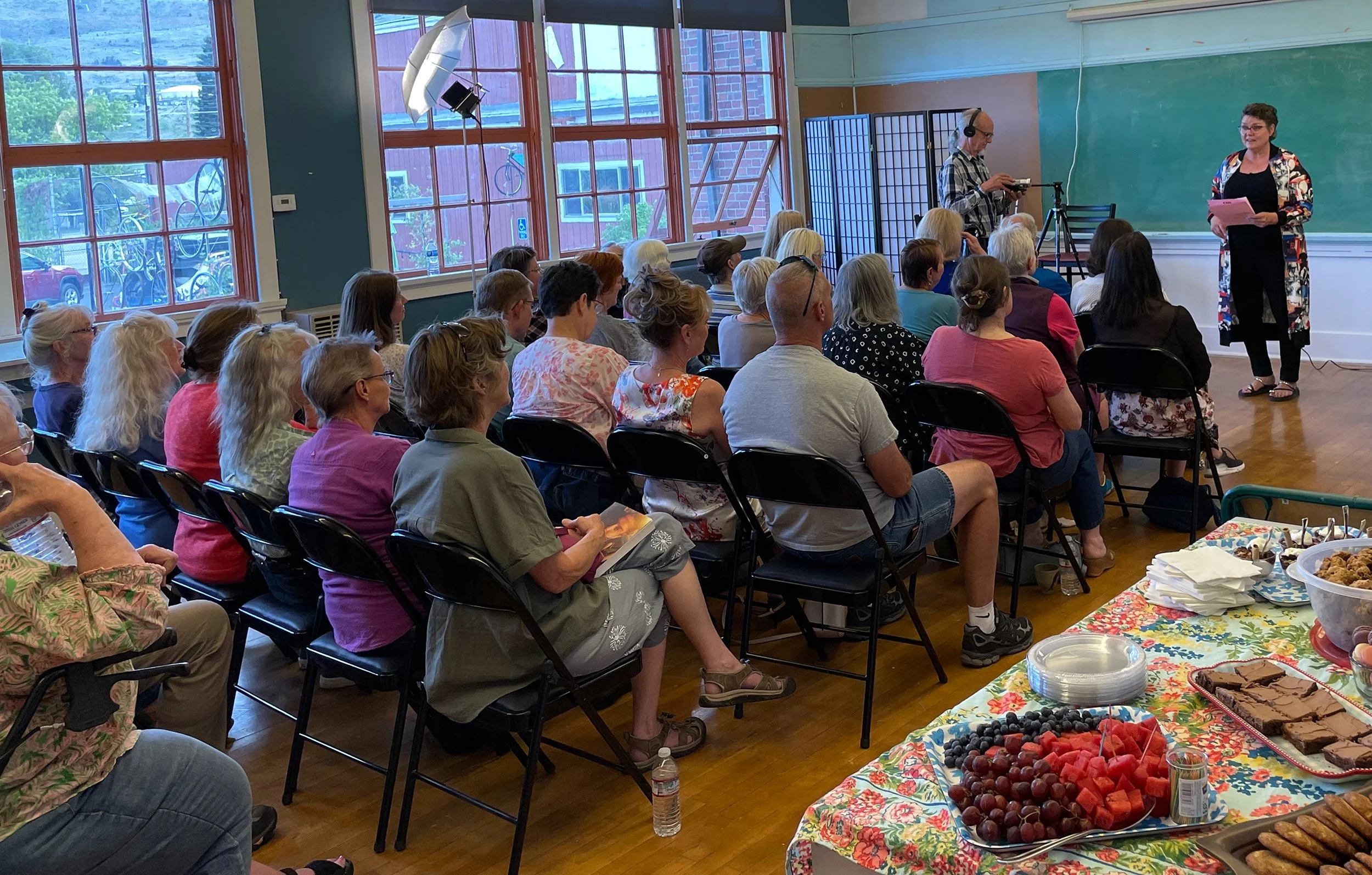 A woman speaking or presenting in front of an audience of adults seated in black chairs in a room with large windows, a chalkboard, and a photographer or videographer capturing the event. A table with snacks and fruit is visible in the foreground.