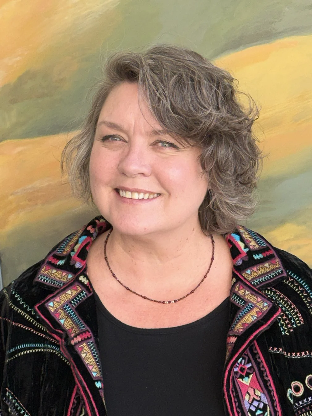 A woman with short, wavy gray hair smiling, wearing a colorful embroidered jacket and a black shirt, standing in front of an abstract painting with green, yellow, and orange hues.