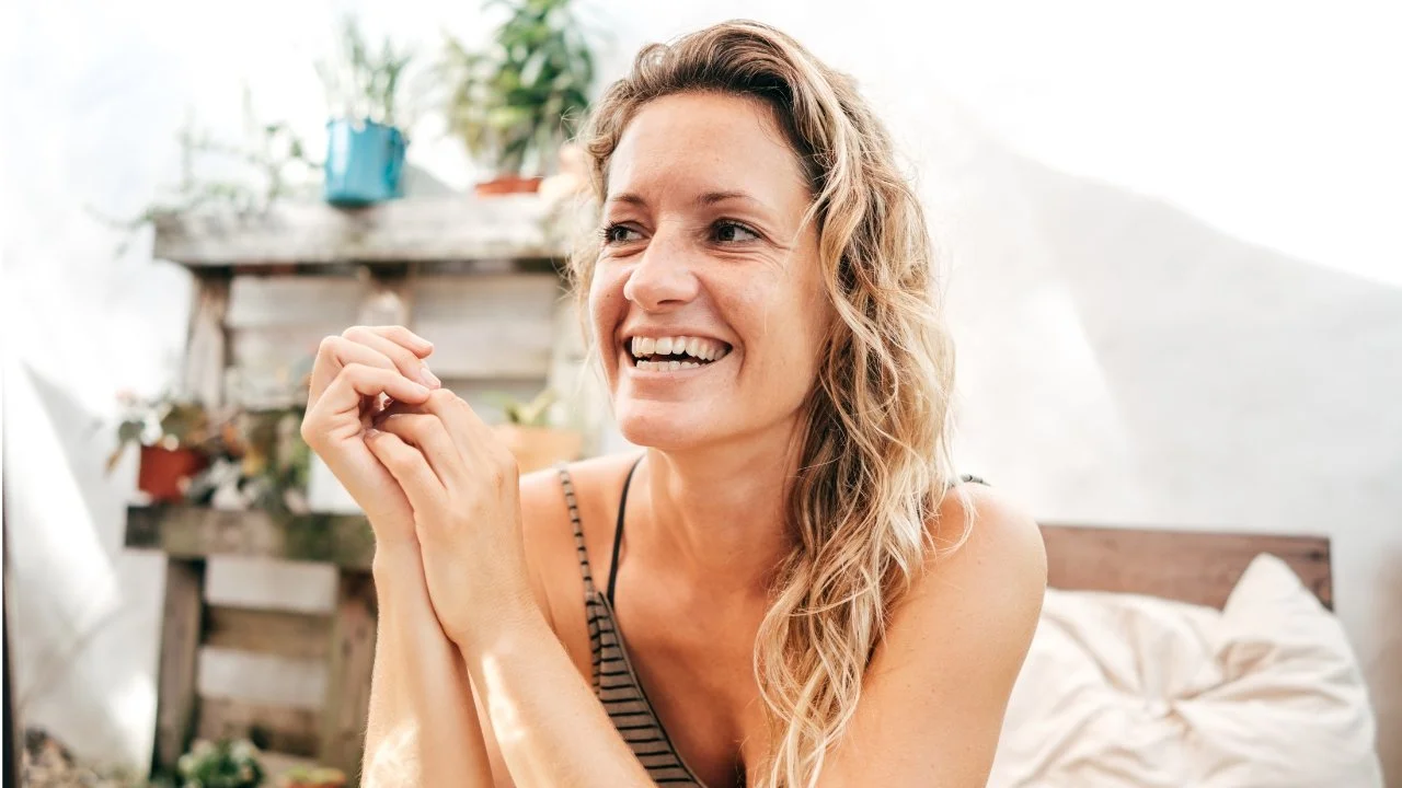 A smiling woman with wavy blonde hair sitting indoors with potted plants and wooden furniture in the background.