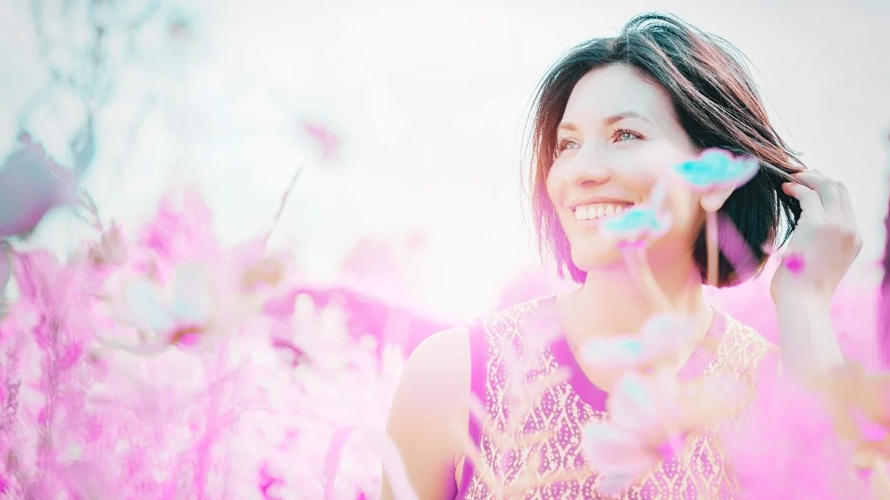 A woman smiling outdoors surrounded by pink and white flowers, with sunlight in the background.