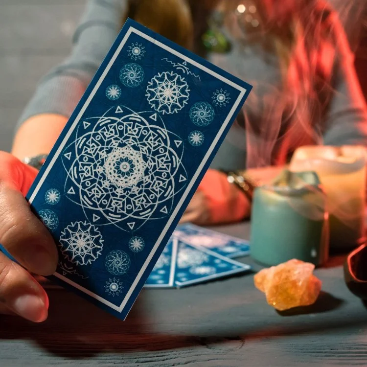 Person holding a blue tarot card with white geometric and snowflake-like designs. In the background, there are other cards, a turquoise mug, a candle, and a yellow stone on a wooden table.