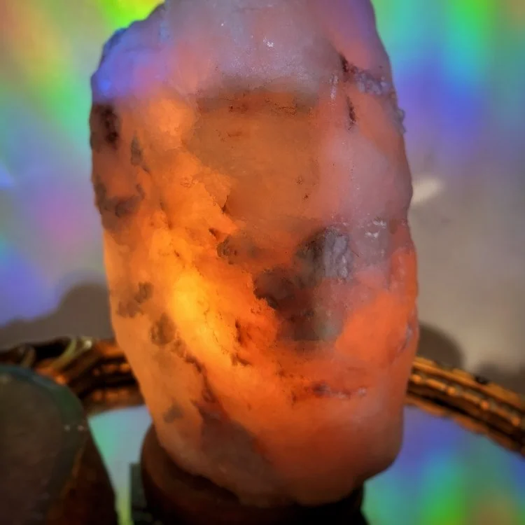 A Himalayan salt lamp shaped like a human face sculpture, illuminated with warm orange light and colorful background reflections.