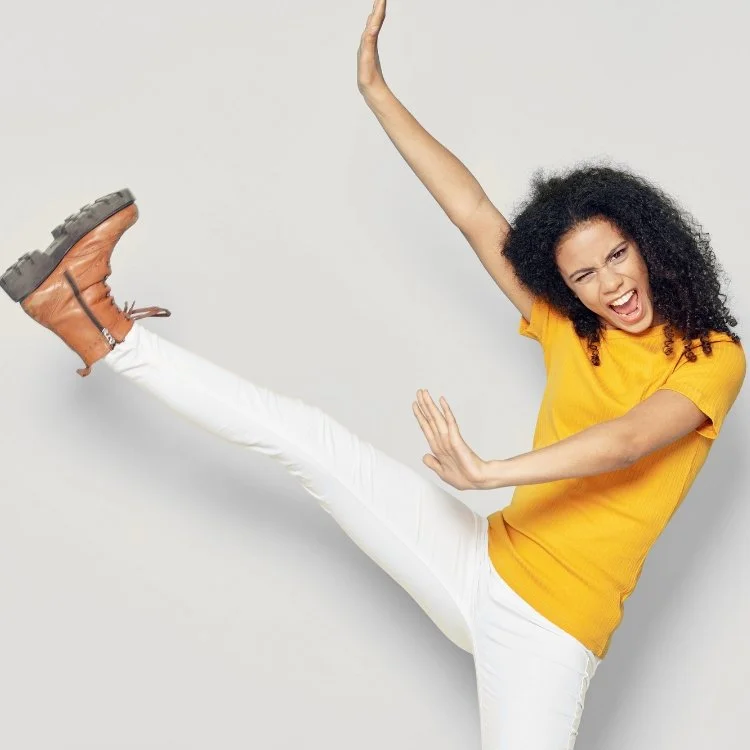 Woman with curly hair wearing a yellow t-shirt and white pants, in a dynamic pose with one leg raised and arms extended, making a playful facial expression.
