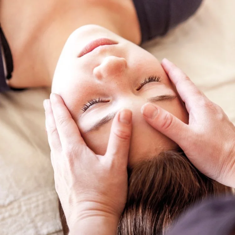 A person receiving a face massage, lying down with closed eyes, with hands gently massaging their forehead.