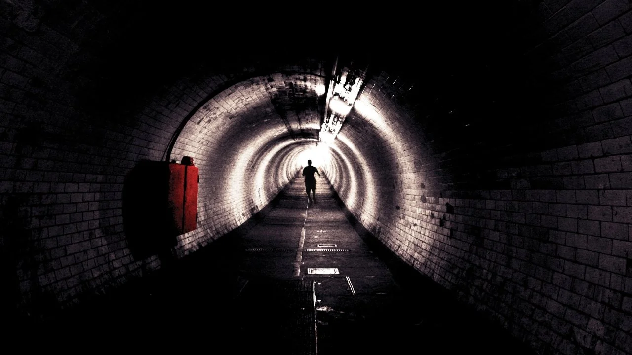 Person walking through a dark, illuminated tunnel with brick walls and a red fire box on the side.