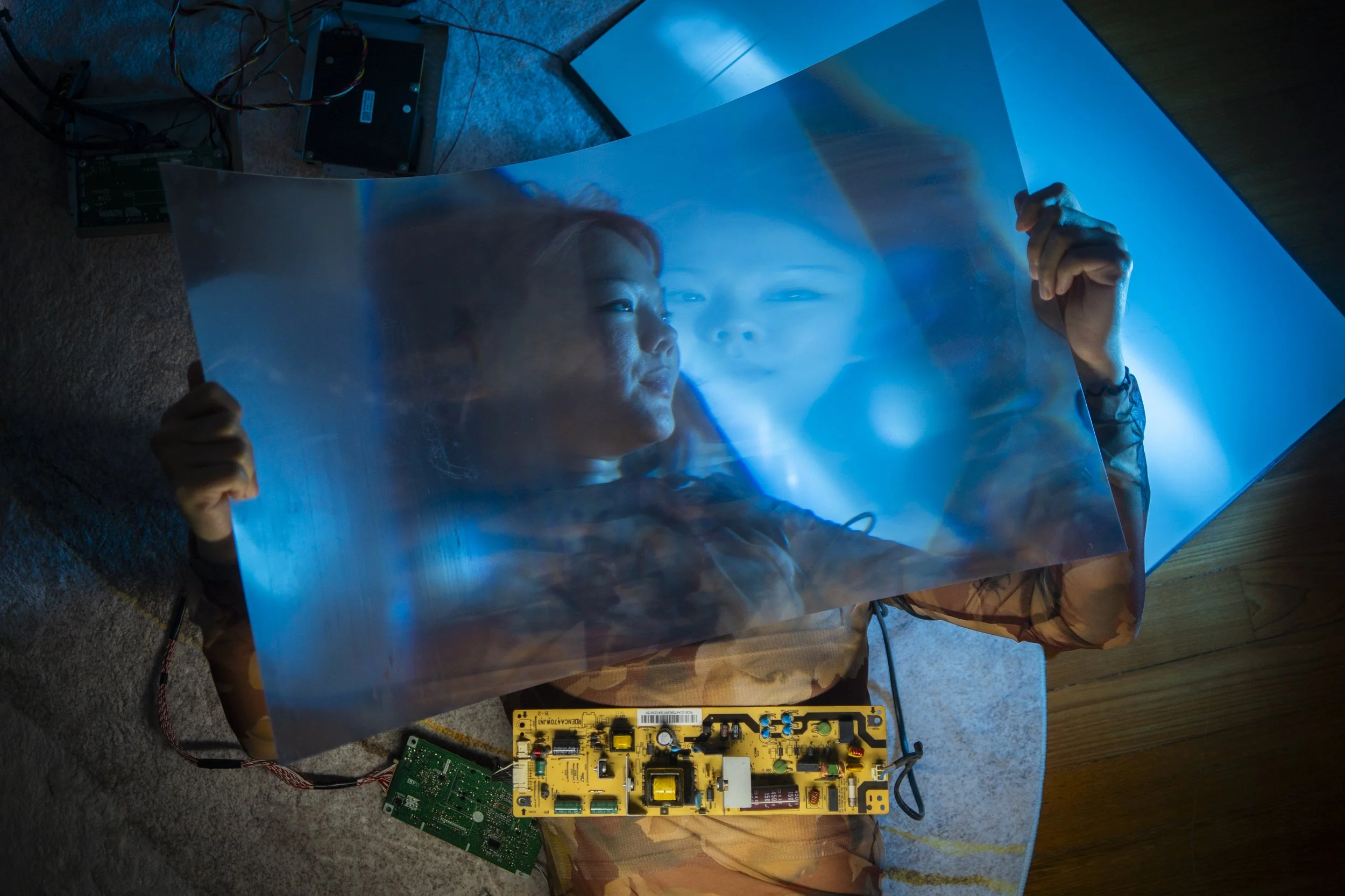 Visual artist Jo Ho, surrounded by circuit boards and wires, holds up a reflective film from a dismantled television as part of her GenAI installation at Objectifs — Centre for Photography and film. (2026)