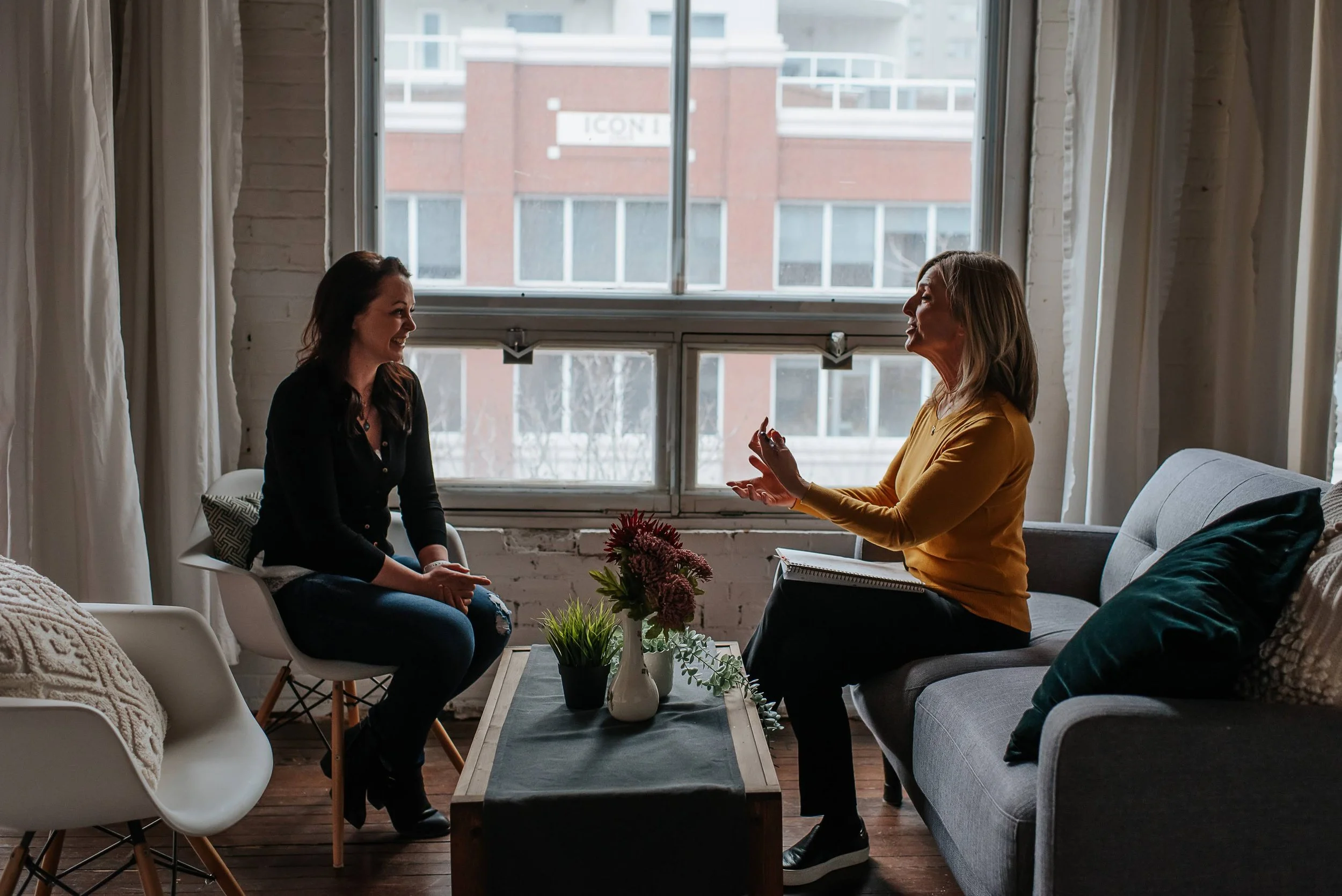 Two women converse in a cozy, modern living room with large window behind them, a small coffee table with flowers in a vase, and casual seating.