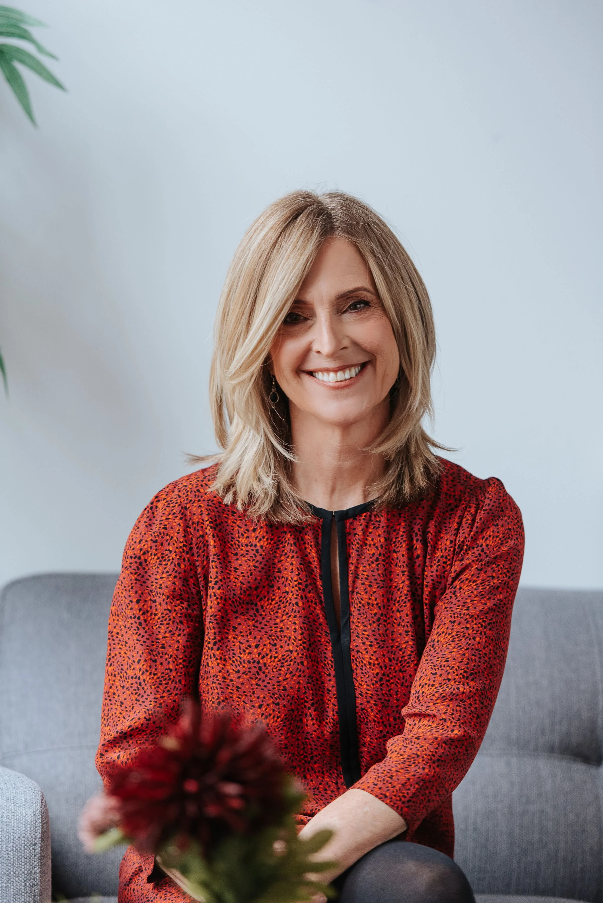 A middle-aged woman with shoulder-length blonde hair, smiling while sitting on a gray couch in a cozy, well-lit room.
