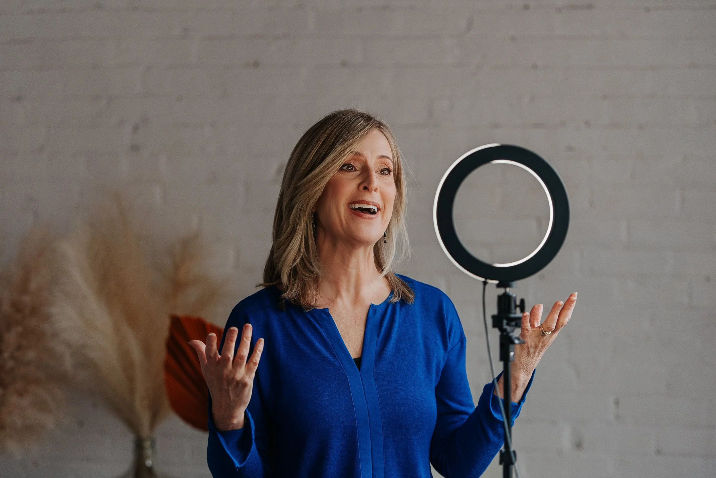 A woman in a blue top speaking in front of a ring light with a white brick wall background.