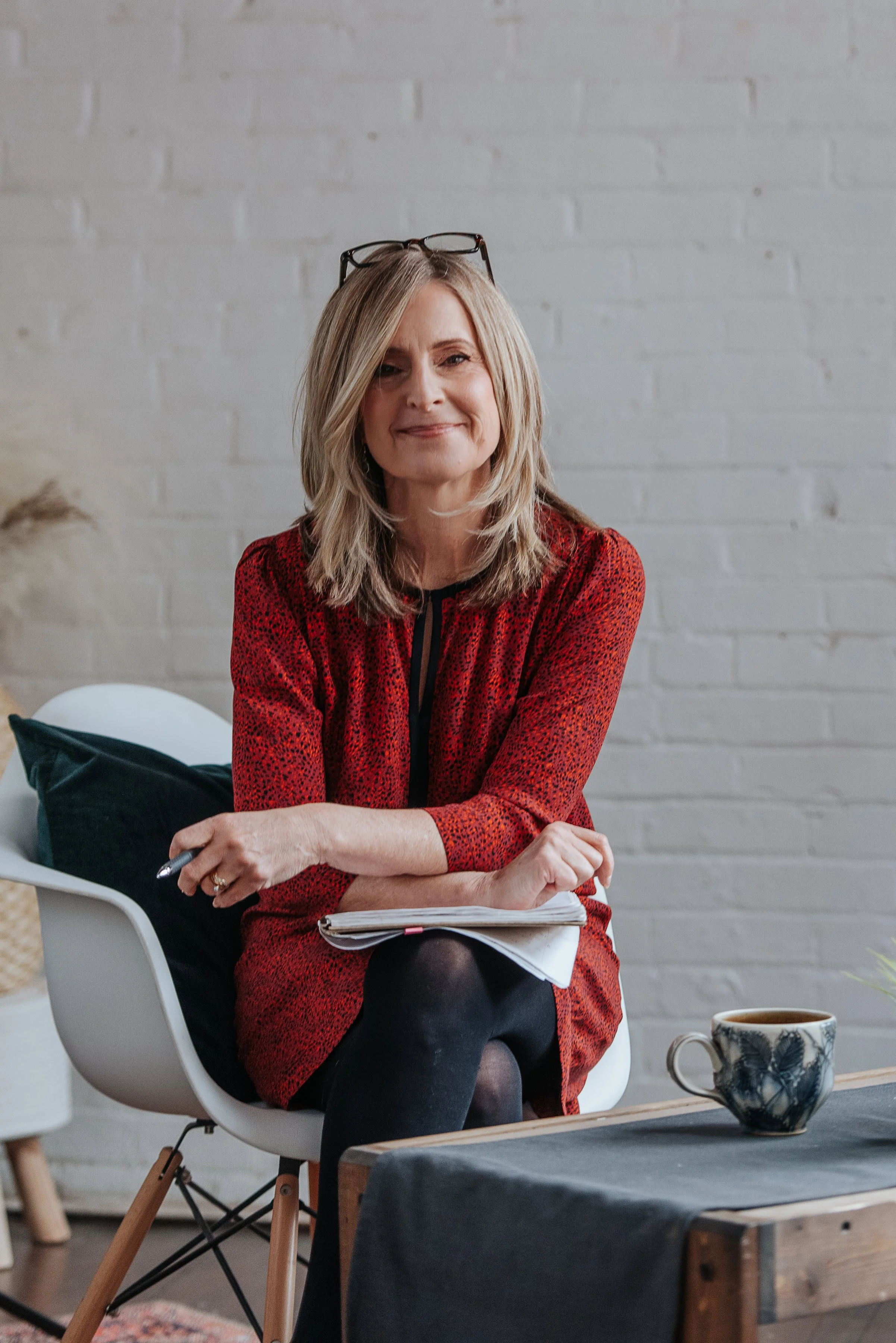 A woman with blonde hair and glasses on her head sitting on a white chair with a notebook in her lap, wearing a red patterned top, in a room with a white brick wall background. A cup on a table in front of her.