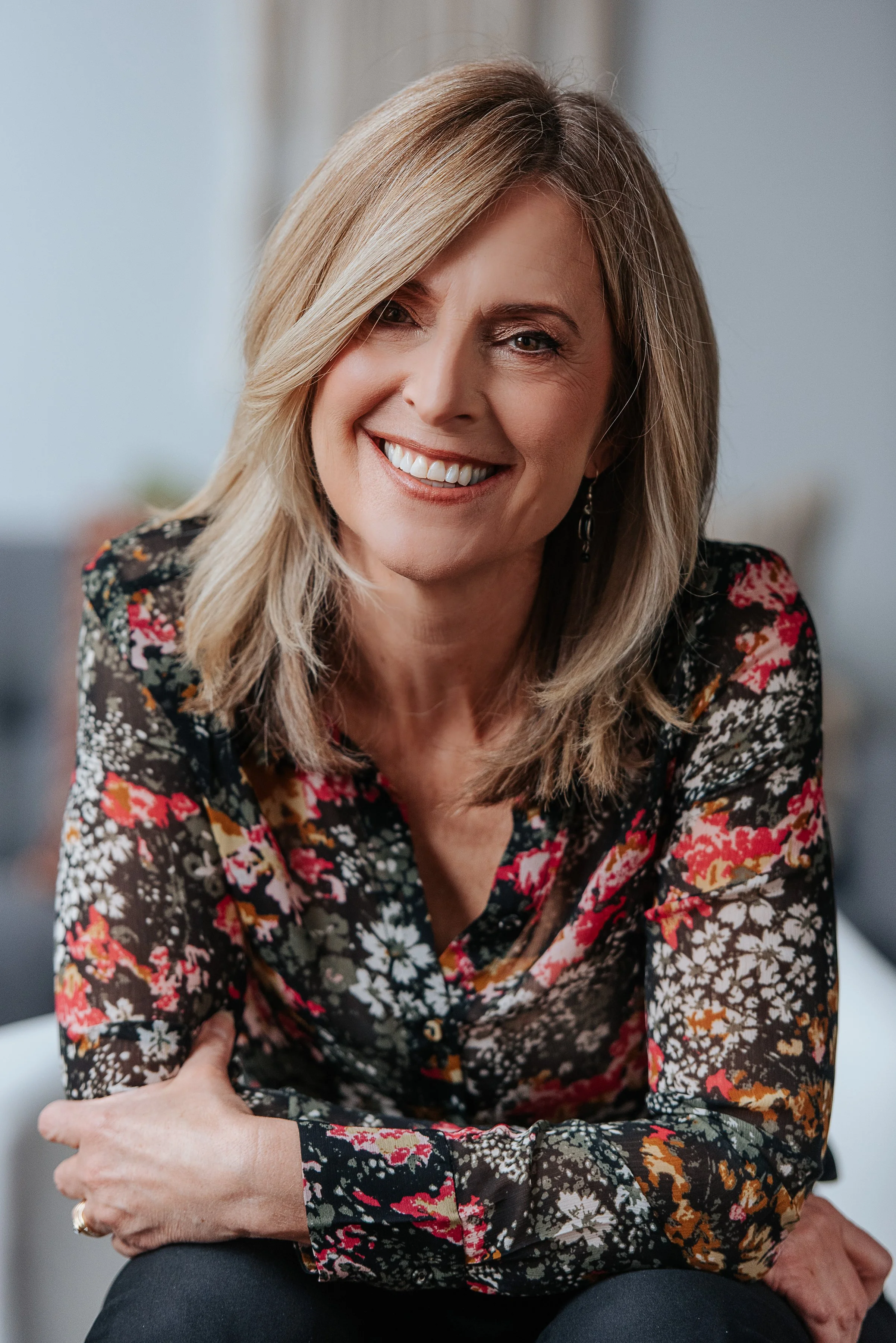 A smiling middle-aged woman with shoulder-length blonde hair, wearing a black floral blouse, sitting indoors.
