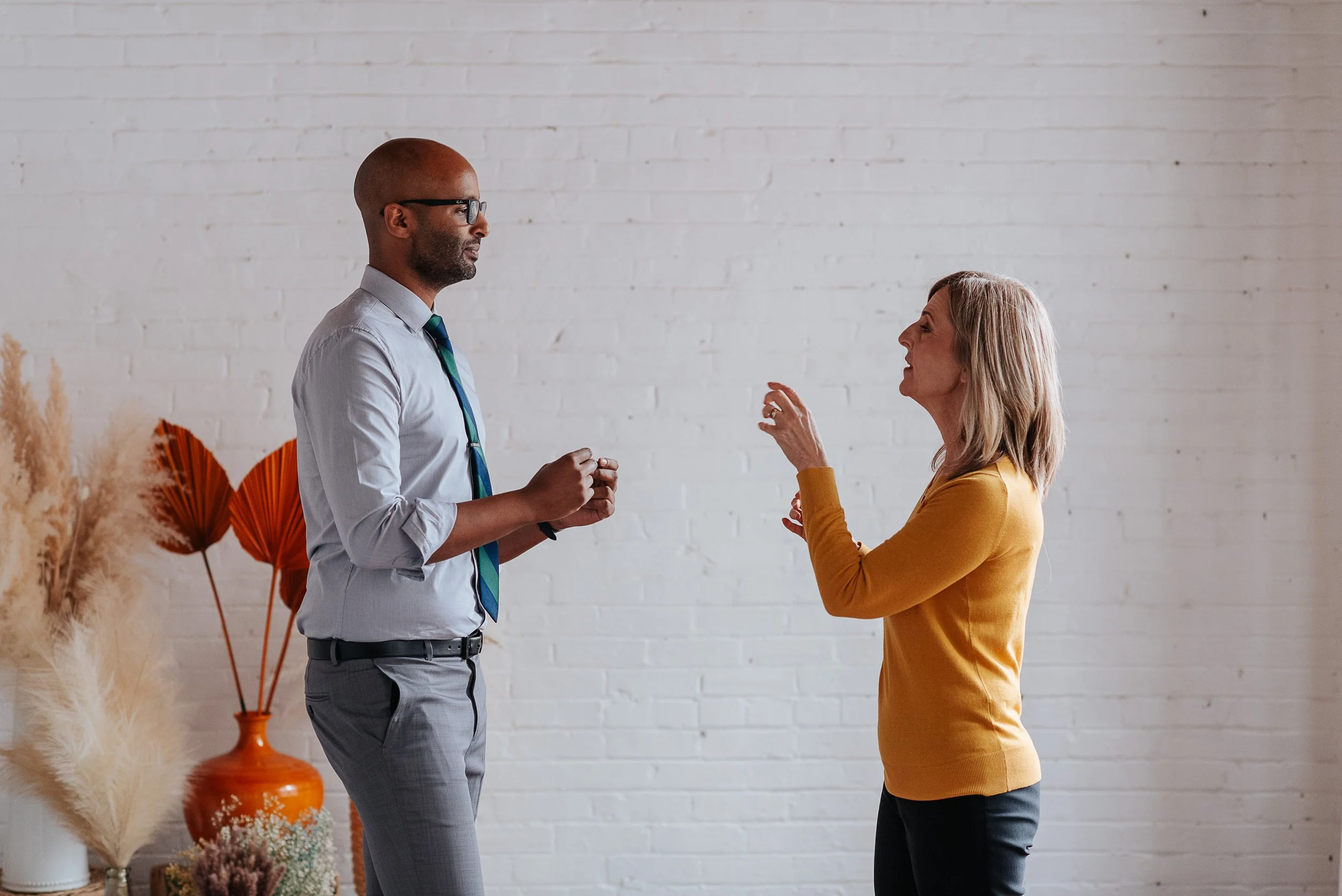 A man and a woman are engaged in conversation indoors, with a white brick wall and decorative plants in the background.