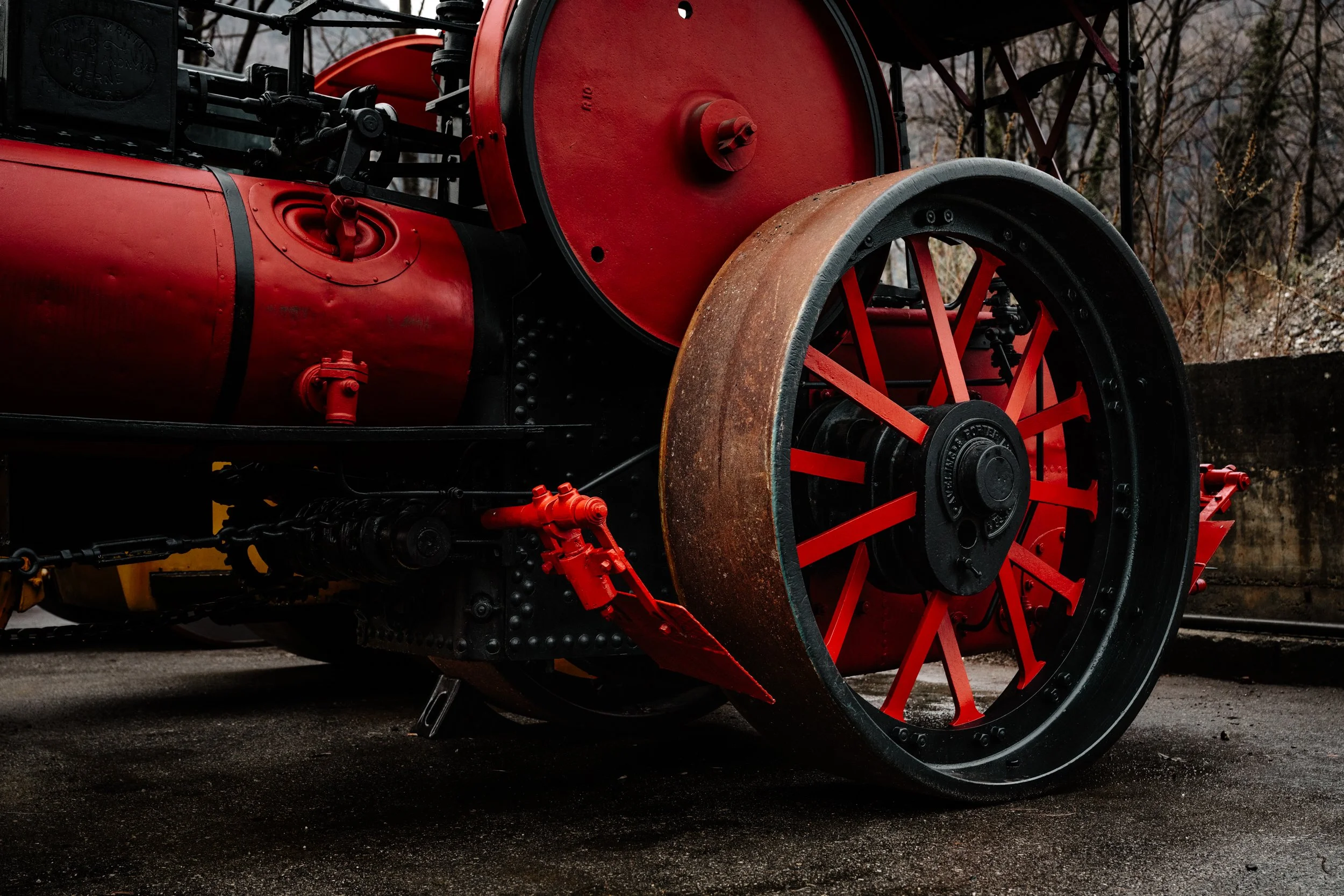 Dettaglio di una locomotiva a vapore rossa, con grandi ruote nere e rosse e altri componenti meccanici visibili, posta su un binario