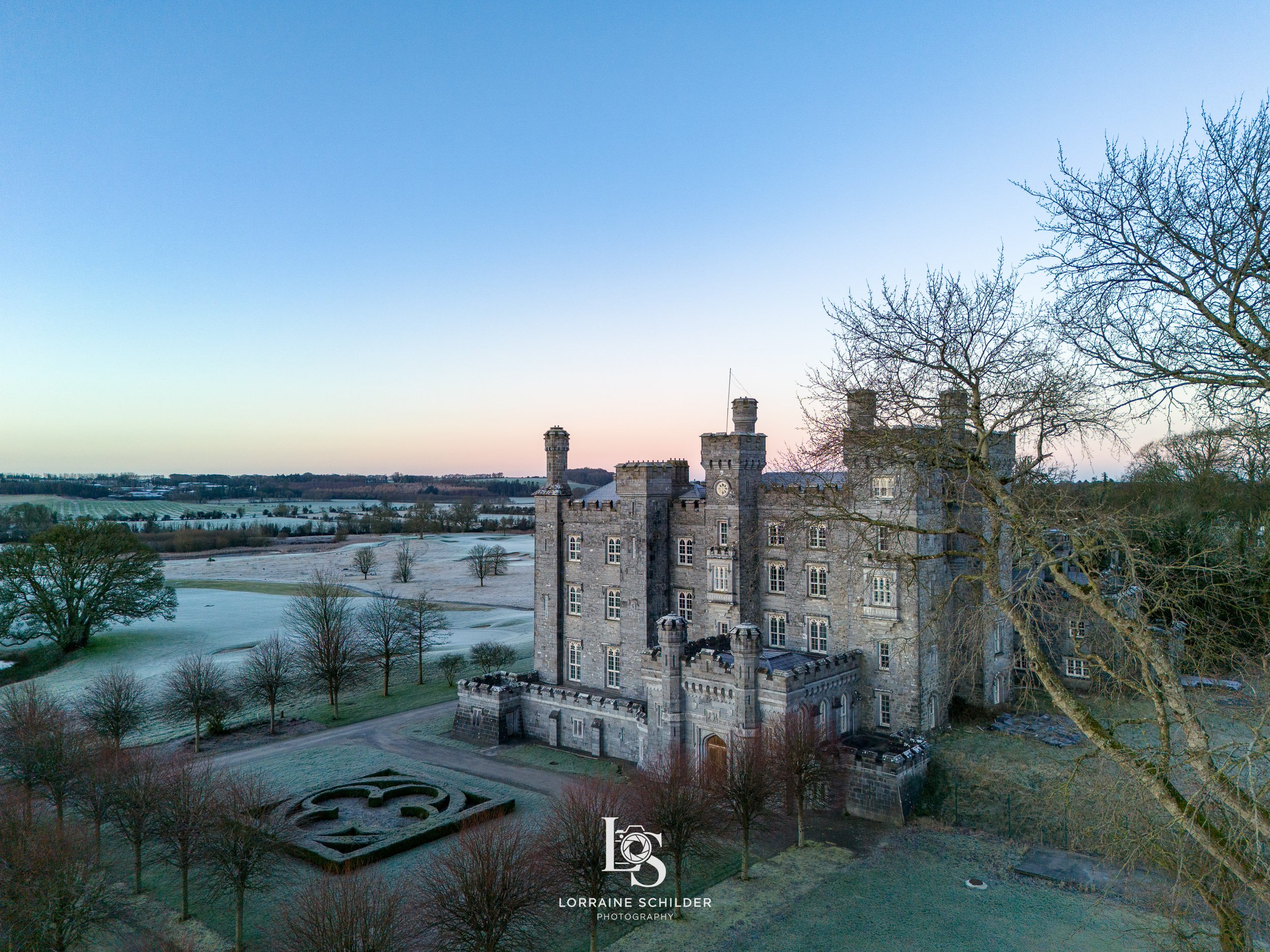 Aerial view of a historic stone castle in a winter landscape with frosted trees and open fields at dusk. Killeen Castle, Meath.