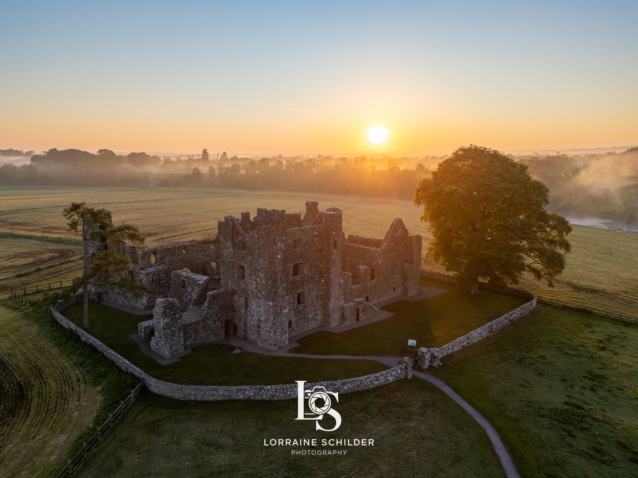 Aerial view of an ancient stone castle surrounded by a stone wall on green grass, with a large tree nearby, at sunrise over a rural landscape with fields and mist.  Bective Abbey, Meath.