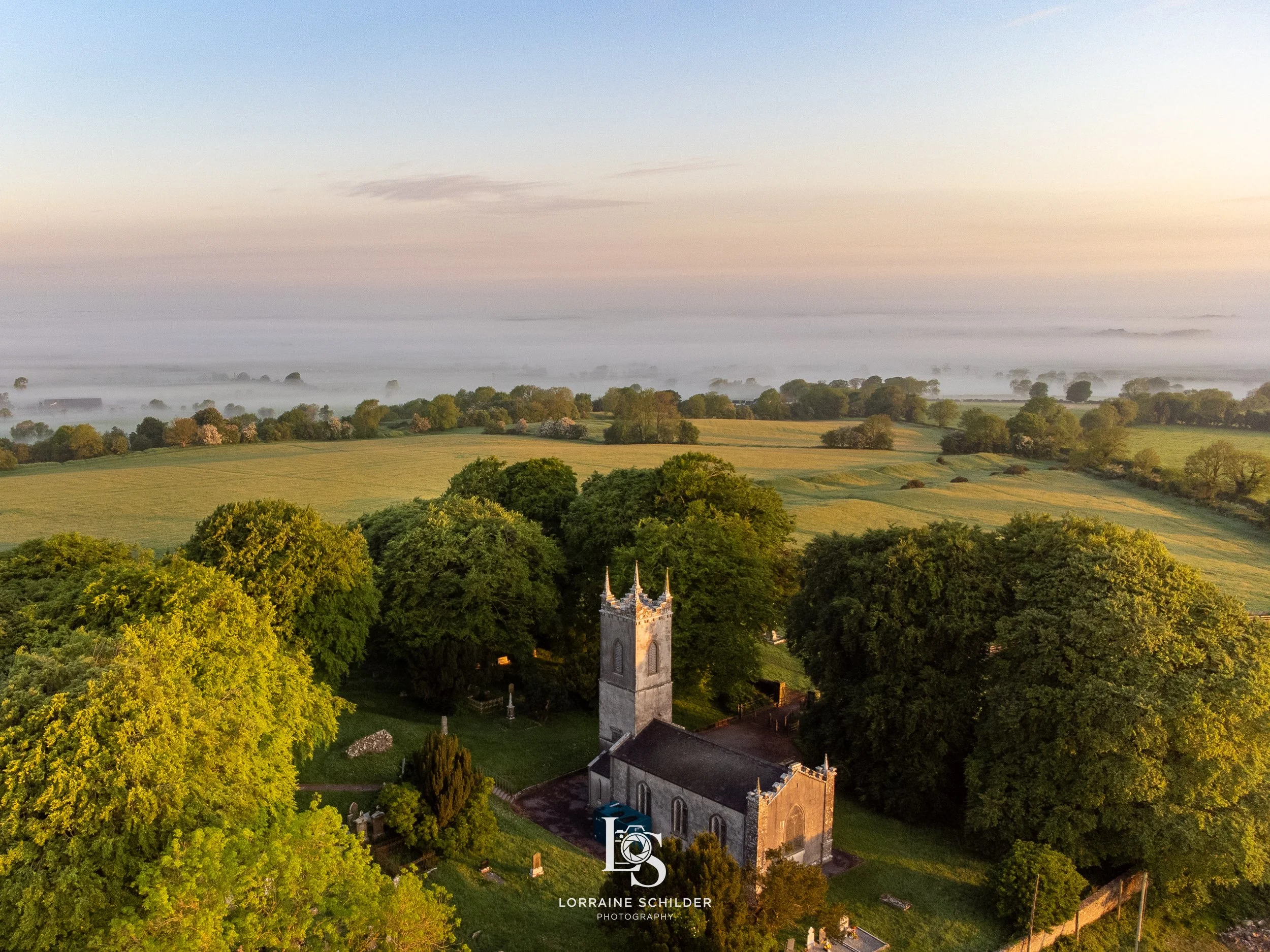Aerial view of a church surrounded by green trees, with rolling fields and a light fog in the distance during sunrise..
