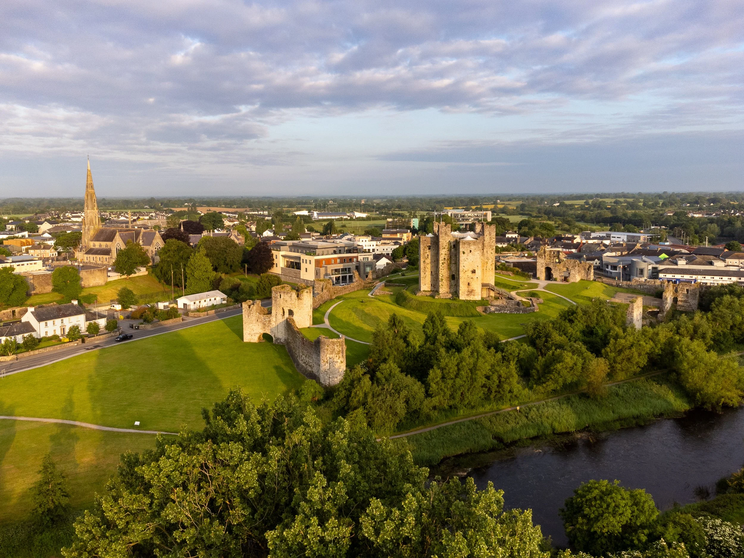 Aerial view of Trim Castle with stone walls and towers, green lawns, a river, and a town with a church spire in the background.  