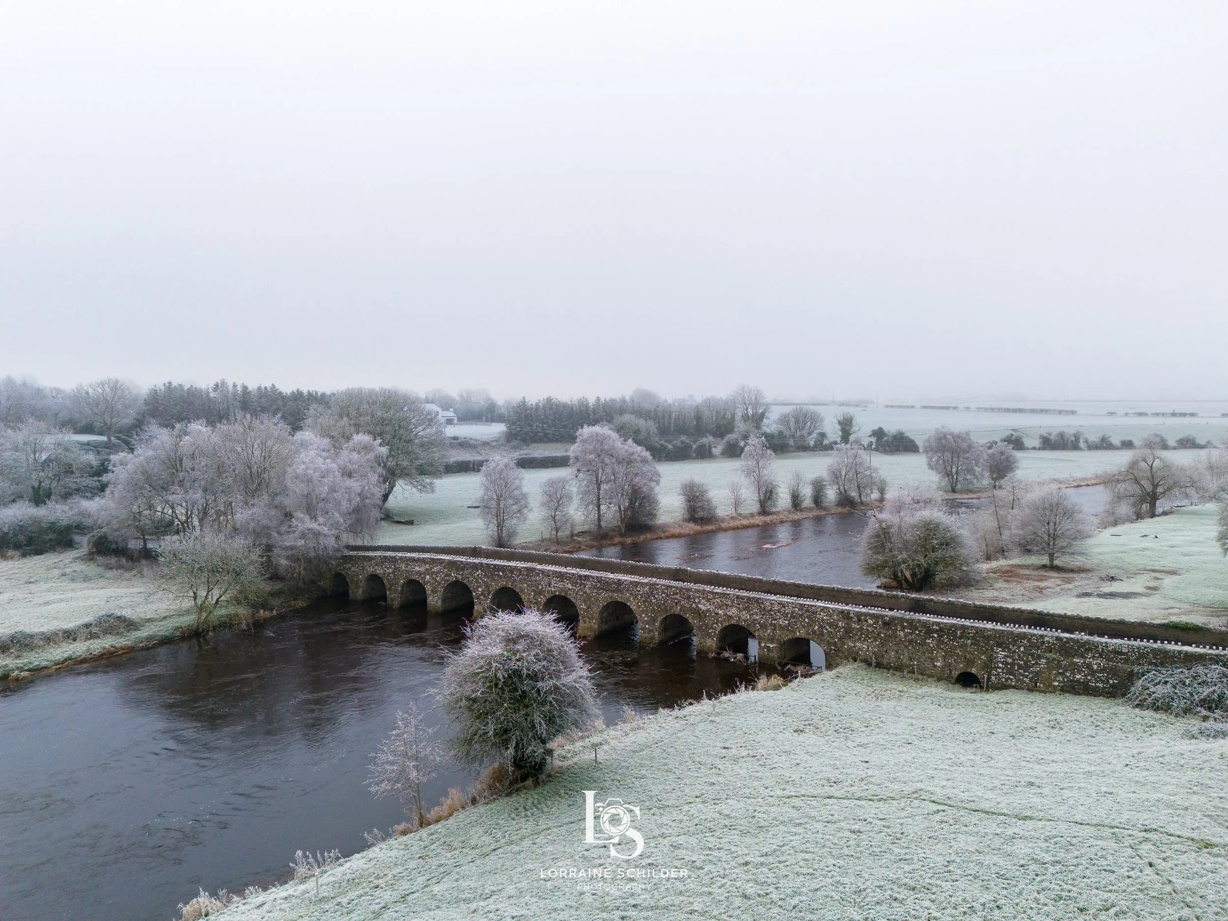 Snow-covered landscape with a river and an arched stone bridge, trees with frosted branches, and a cloudy sky. Bective Bridge, Bective Abbey, Meath.