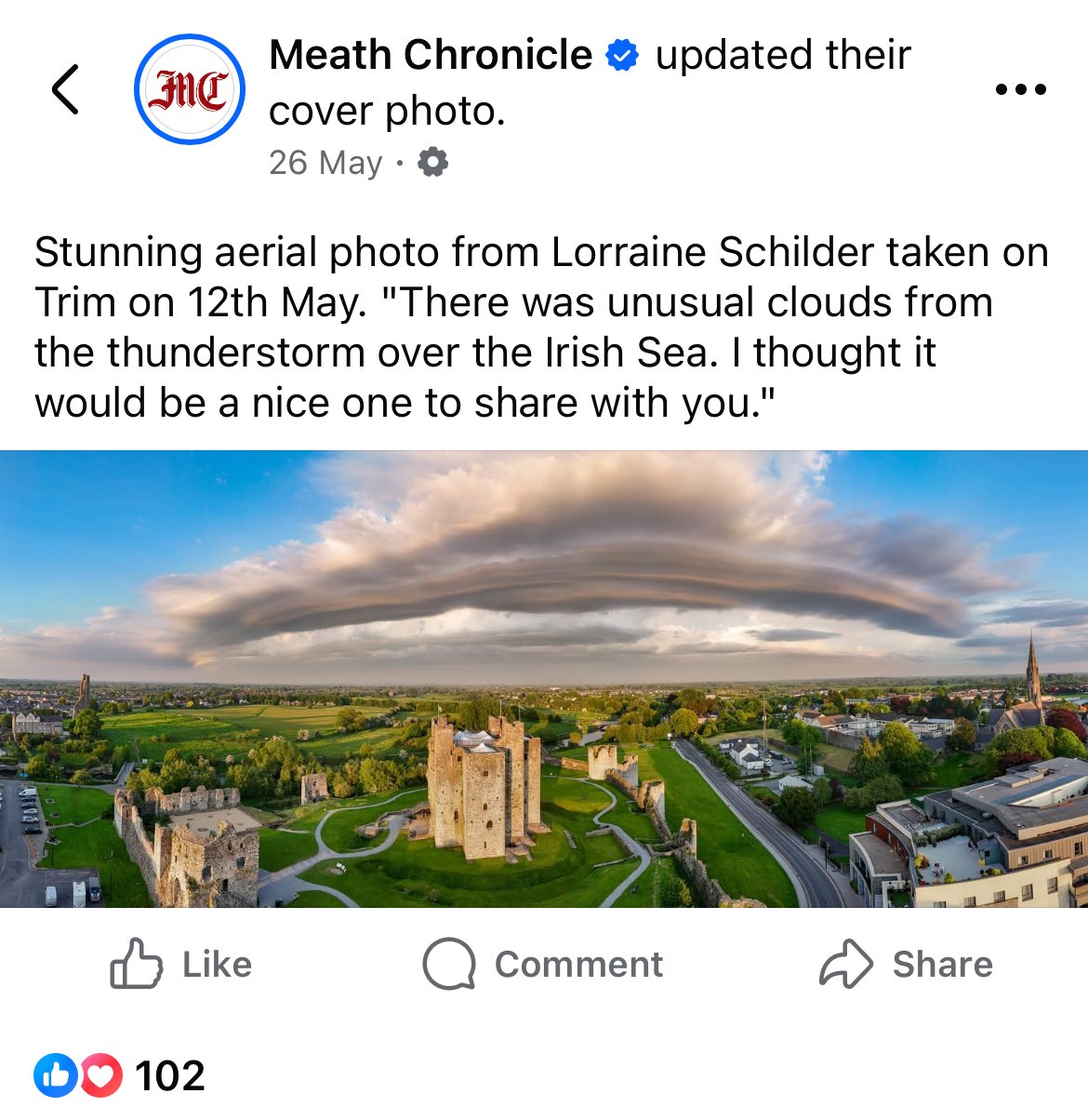 Aerial view of a historic castle with surrounding greenery under a dramatic cloud formation over a town.