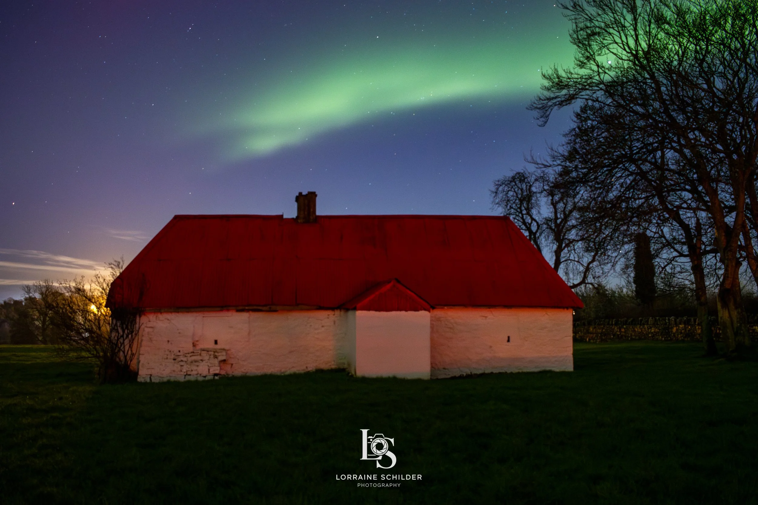 Nighttime scene of a small white house with a red roof, surrounded by leafless trees, under a sky illuminated by green northern lights.