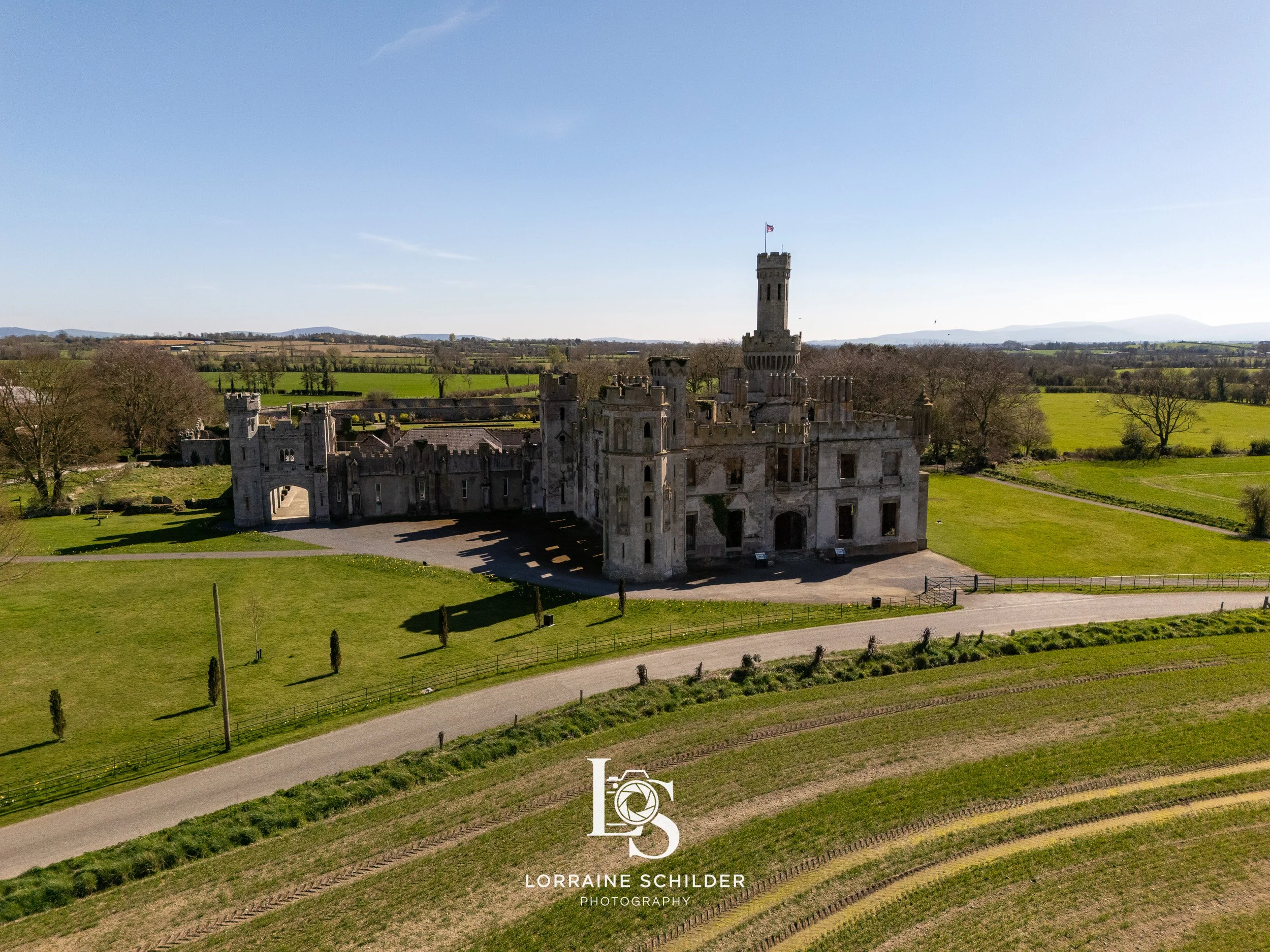 Aerial view of a historic stone castle with towers and a courtyard, surrounded by green fields and trees under a clear blue sky. Carlow.