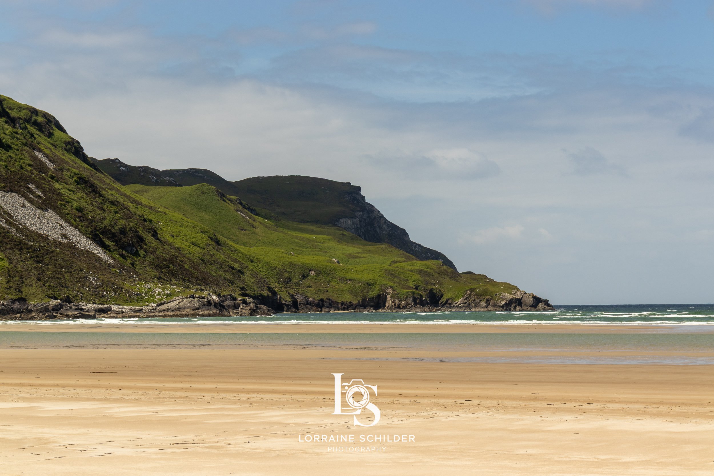 A serene beach with golden sand in the foreground, green rolling hills and cliffs in the background, and a partly cloudy sky overhead.  Donegal.
