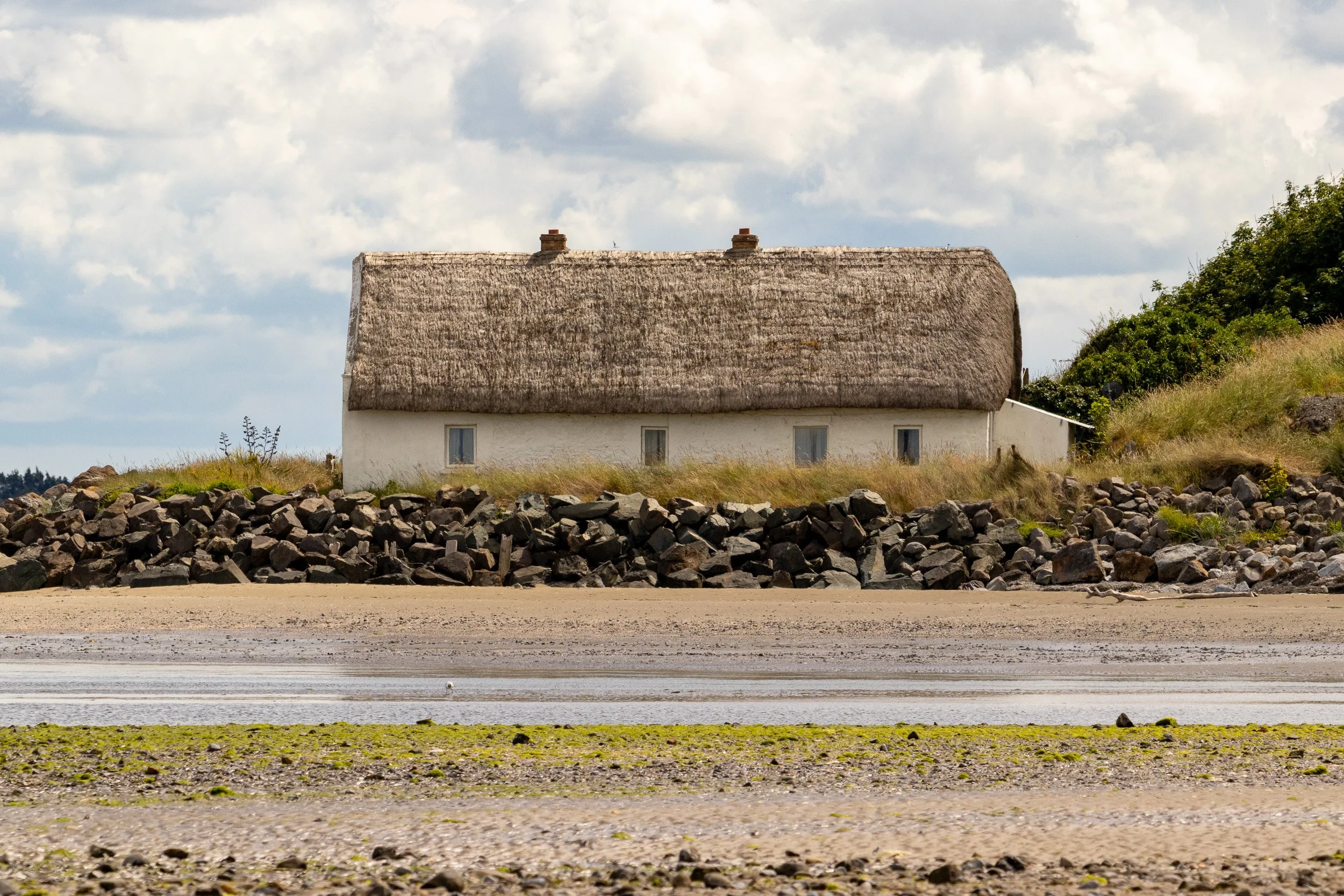 A white house with a thatched roof on a grassy hill, rocky embankment, sandy shore, and cloudy sky.