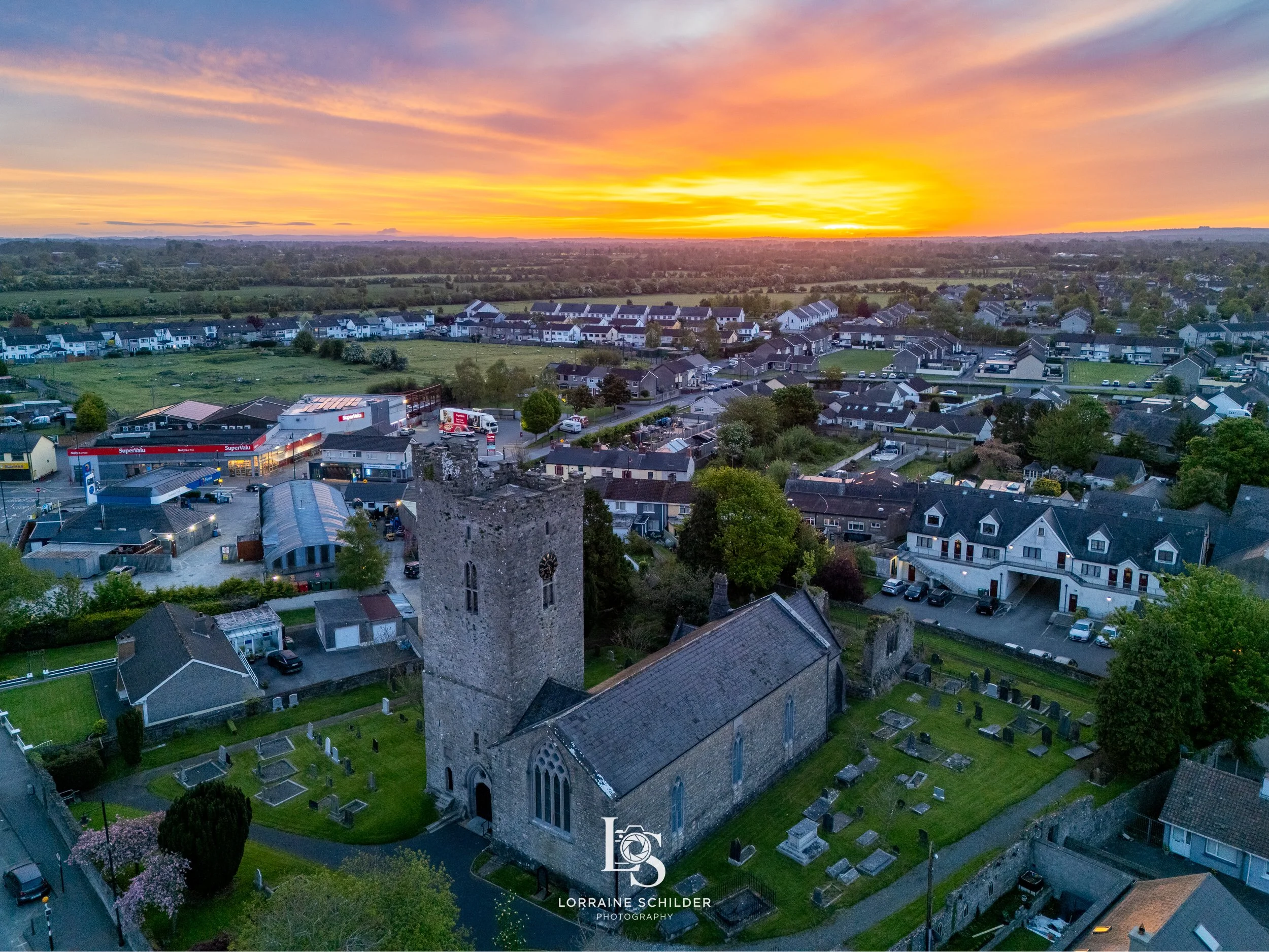 An aerial view of Trim at sunrise, featuring St. Patricks Cathedral,  surrounding graveyard, modern residential homes, and commercial buildings, with a vibrant orange sky and green fields in the distance.