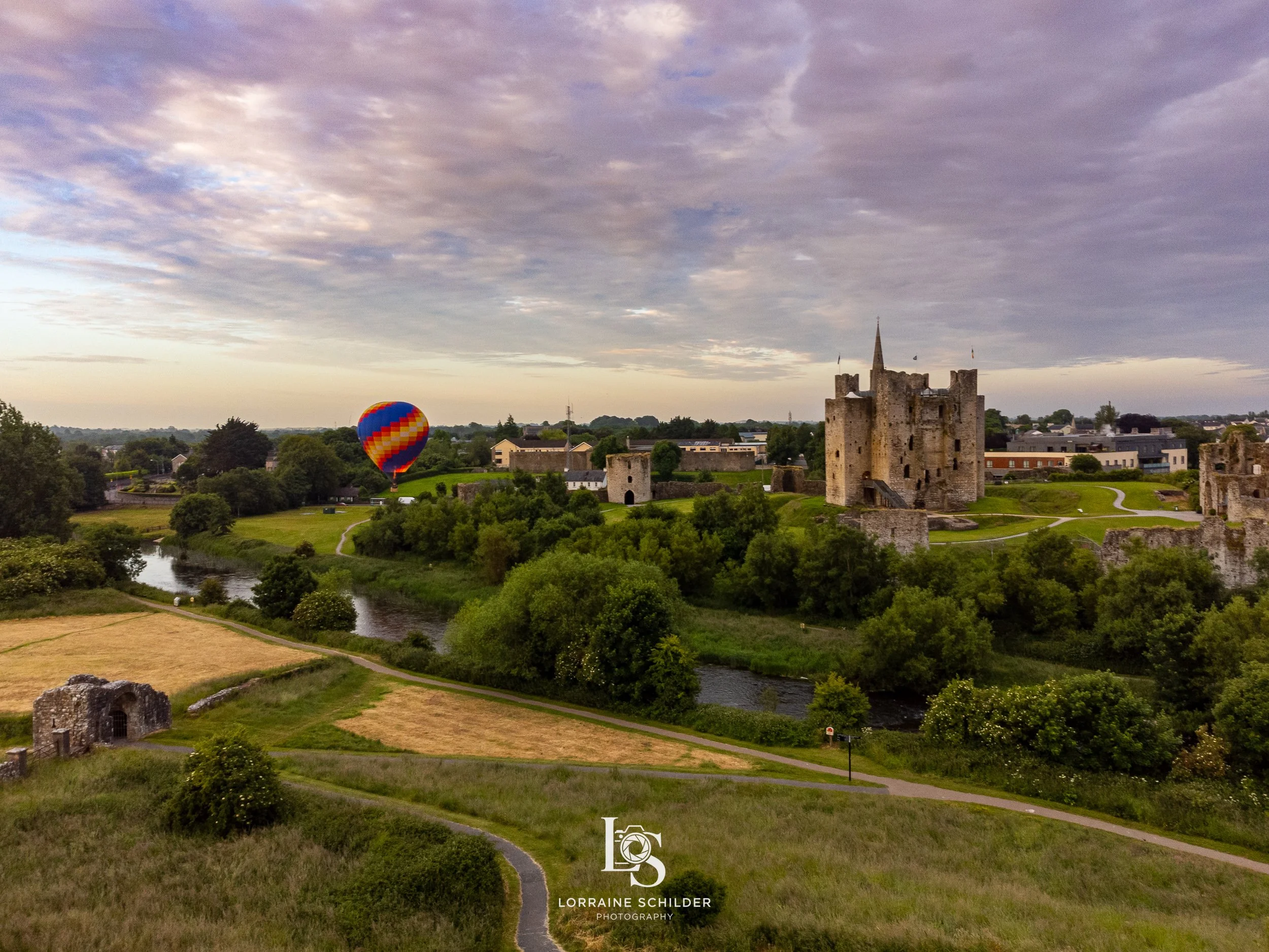 A scenic landscape of Trim castle surrounded by greenery and a river, with a colorful hot air balloon floating in the sky at sunrise.