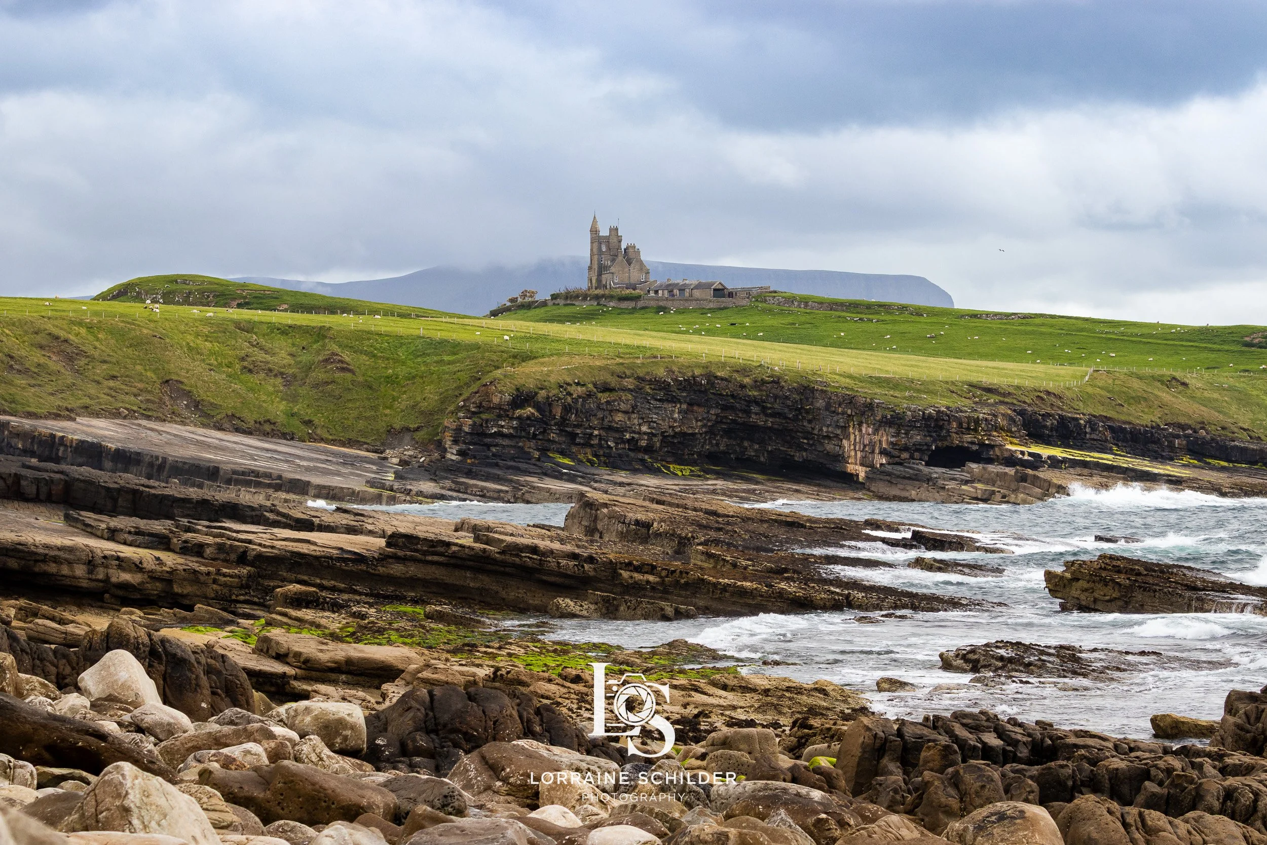 A coastal landscape with rocky shoreline in the foreground, green grassy hills and cliffs in the midground, and a historic castle on a hilltop in the background, under a cloudy sky. Sligo.