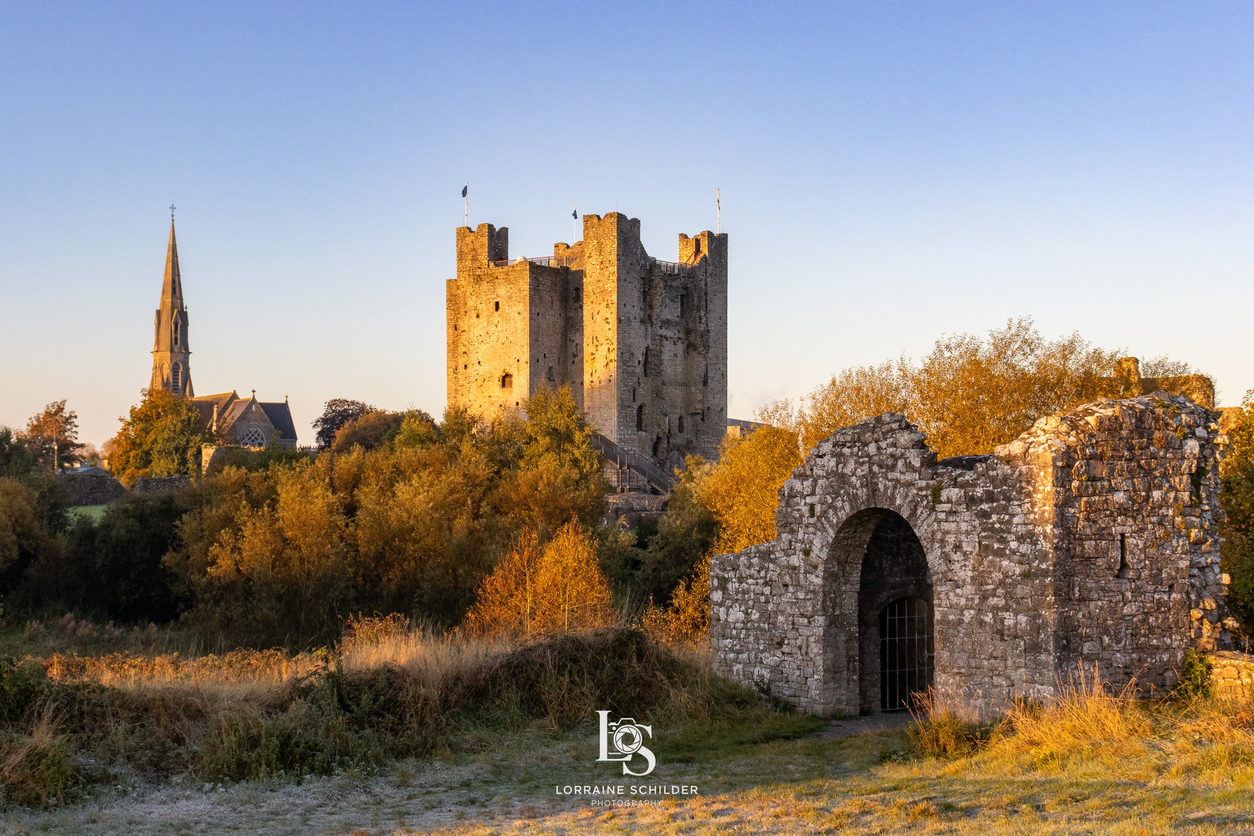 Trim Castle with stone walls and a tall church tower, surrounded by trees with autumn foliage, and a distant church steeple under a clear blue sky at sunrise.