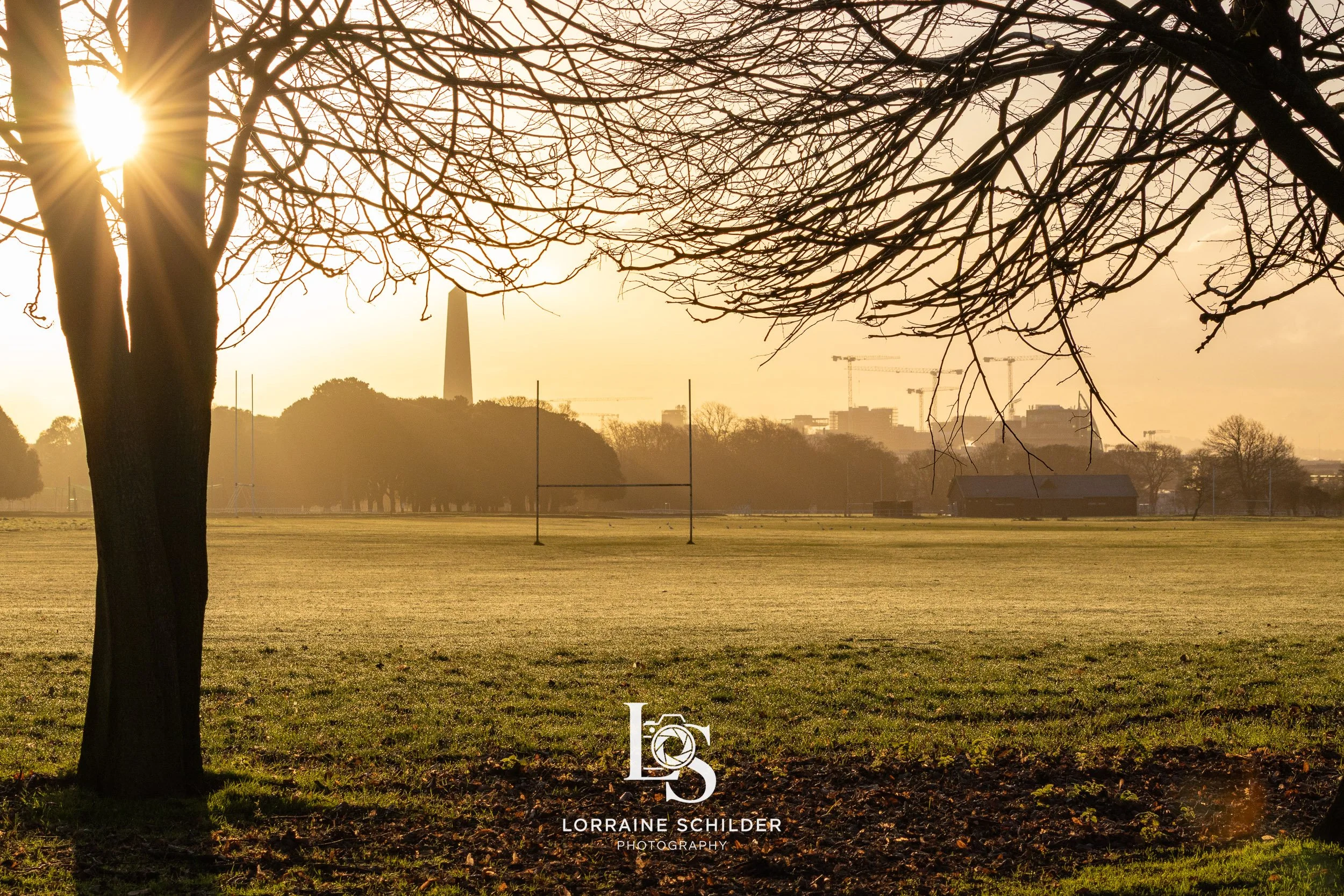 A scenic outdoor view of a park during sunrise  with leafless trees, open grass field, goalposts, and distant industrial buildings with cranes in the background. Dublin, Ireland
