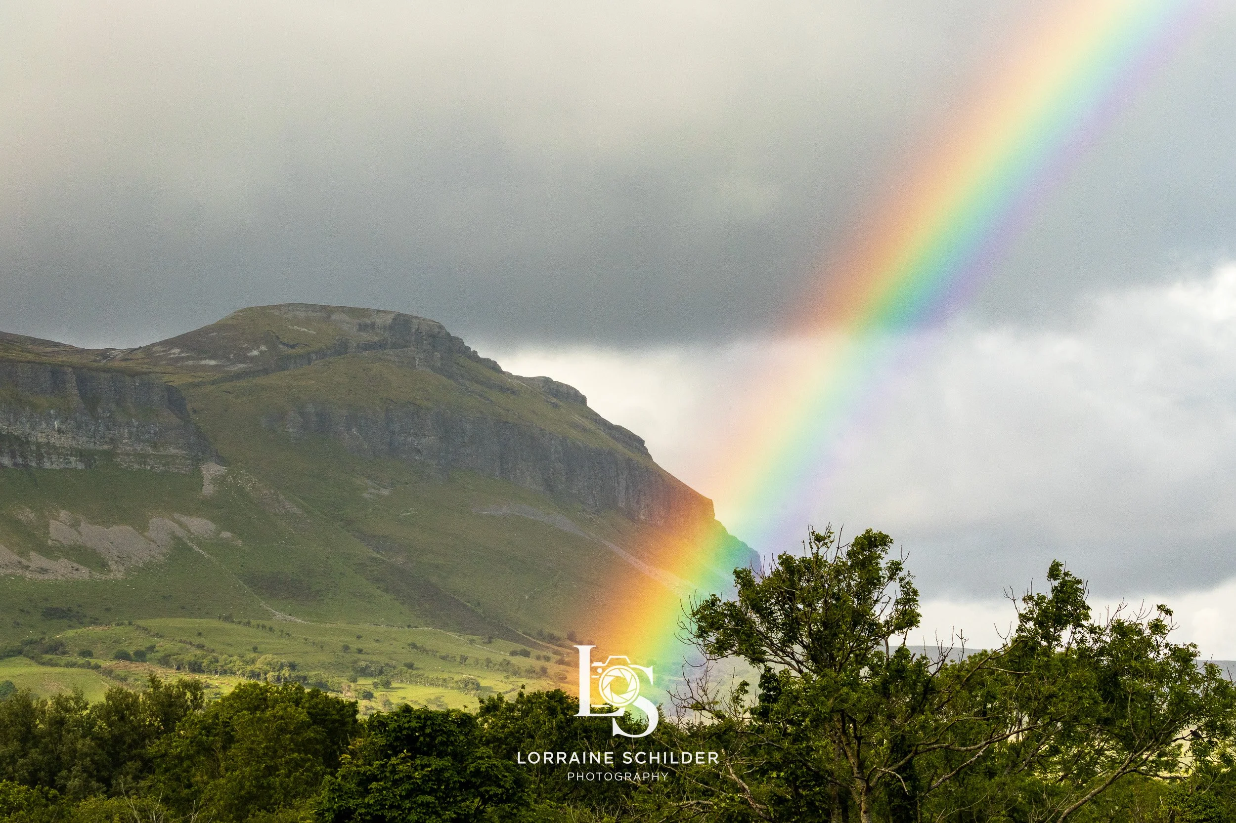 Rainbow arching over a green hillside with trees in the foreground and a large mountain in the background, under a cloudy sky. Sligo.