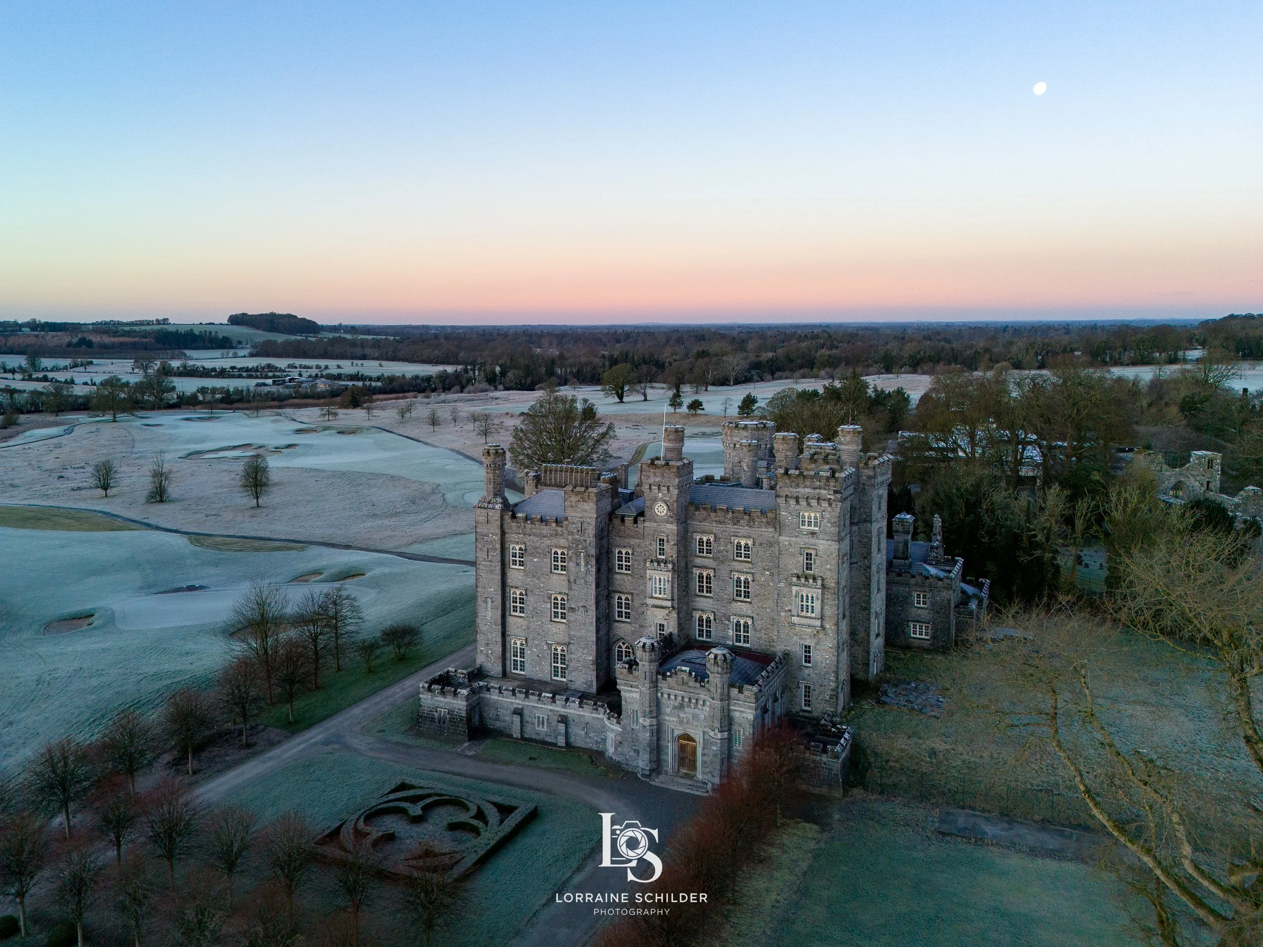 An aerial view of a large stone castle surrounded by trees and open fields during early morning or late afternoon, with a visible moon in the sky. Killeen Castle, Meath.