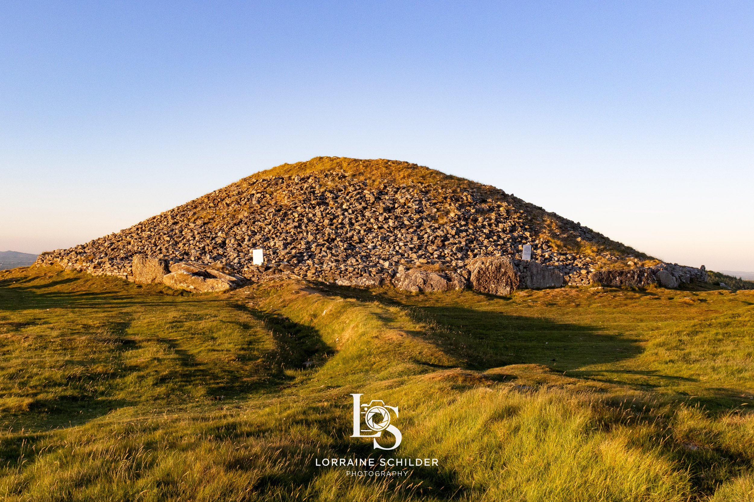 A rocky hill or mound with grass in the foreground, illuminated by sunlight during sunset, with a clear sky in the background. Loughcrew, Meath.