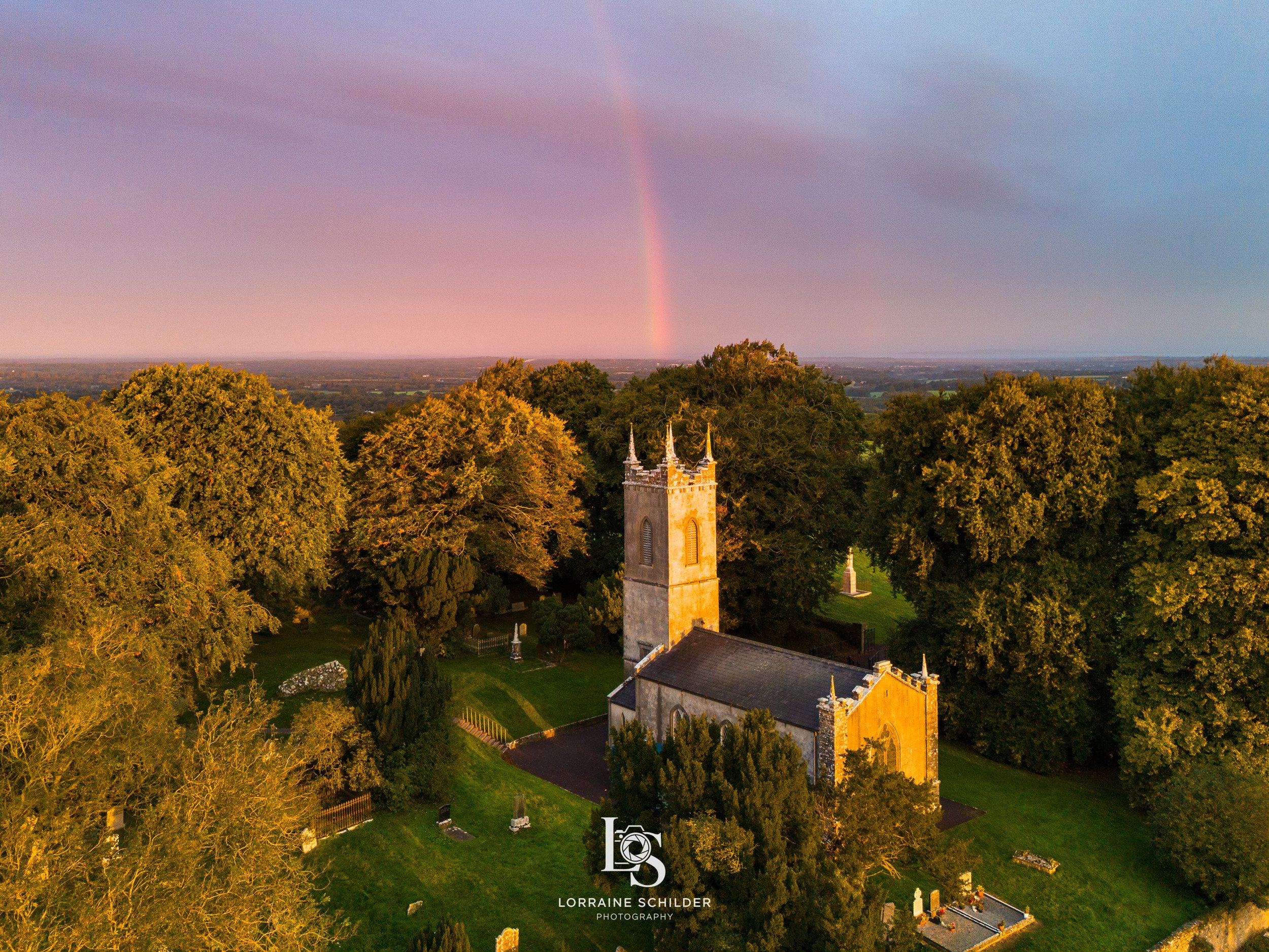 A church surrounded by trees with a rainbow in the sky during sunrise.