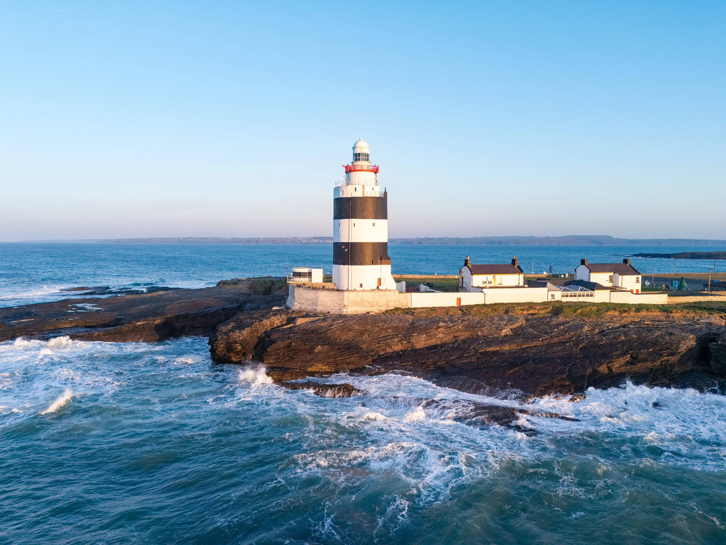 A black and white striped lighthouse on a rocky coastline with waves crashing at the shore, under a clear blue sky.